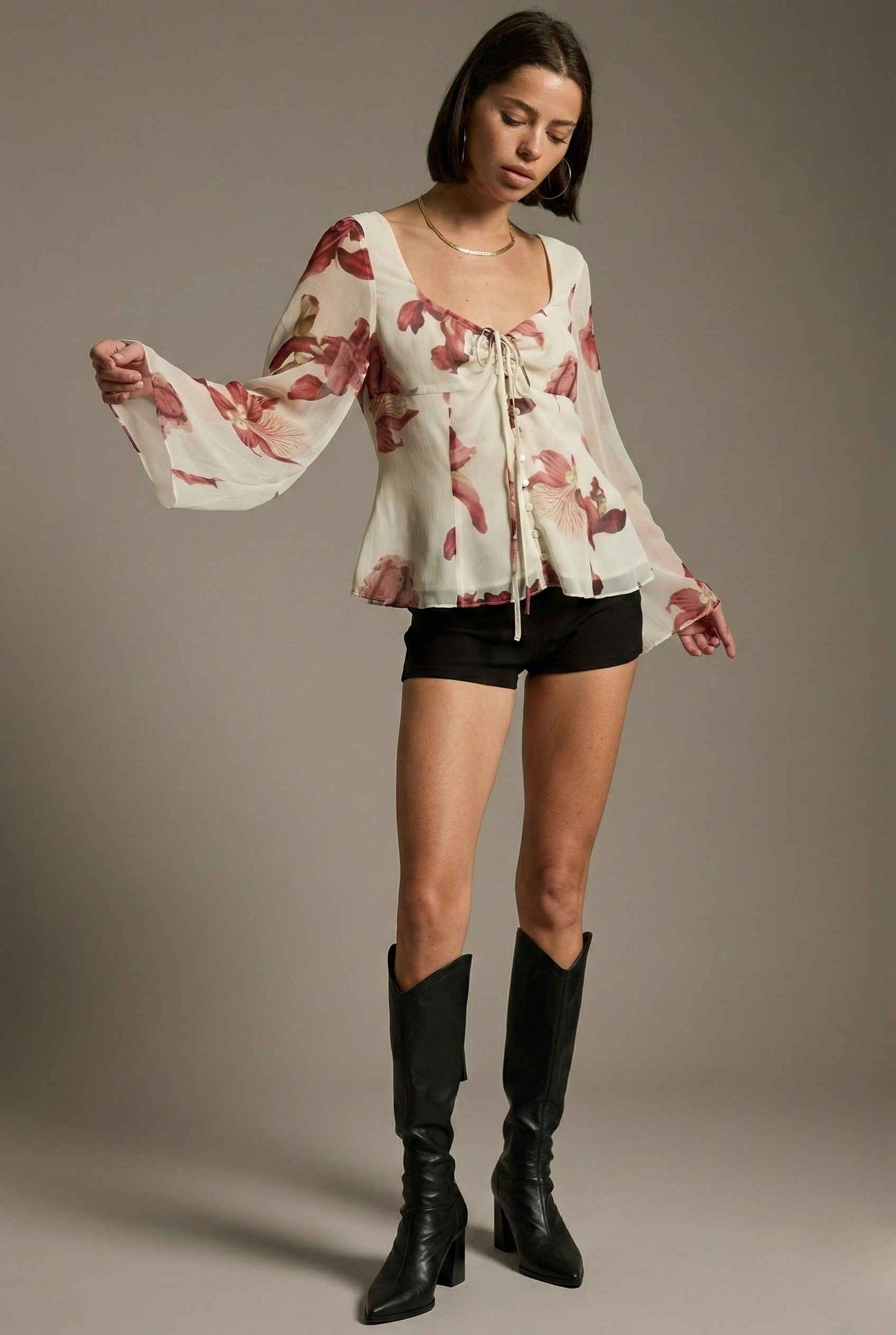 Woman wearing a Aria Top Ivory Floral Tie-Front Bell Sleeve Blouse, black shorts, and black knee-high boots against a plain background