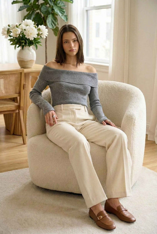 Model wearing Aria Top Ribbed Knit Off-Shoulder Sweater with Side Zipper in Grey paired with trousers and loafers sitting in luxury living room