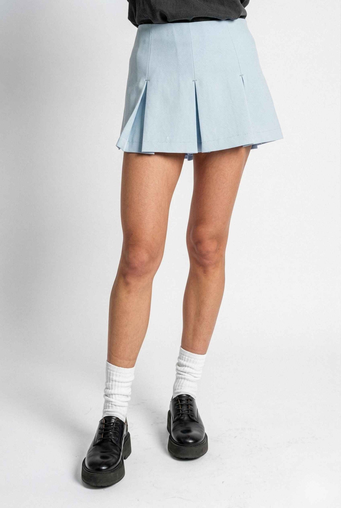 Woman wearing a Celeste Skort Baby Blue Pleated Silk-Blend Mini Skort with black shoes and white socks on a plain background