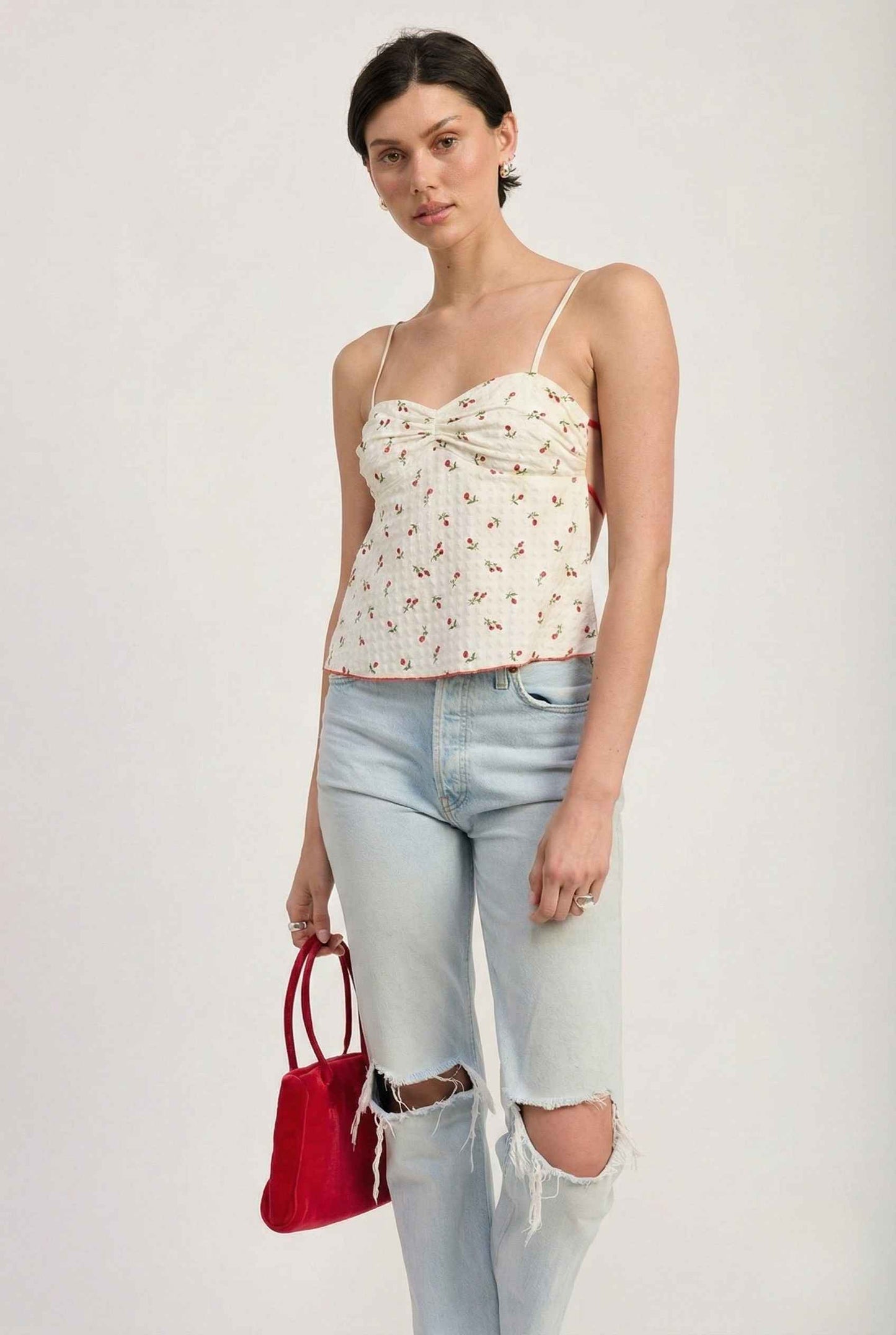Woman wearing a Chloe Top White & Red Floral Sweetheart Camisole and ripped jeans with a red handbag on a white background