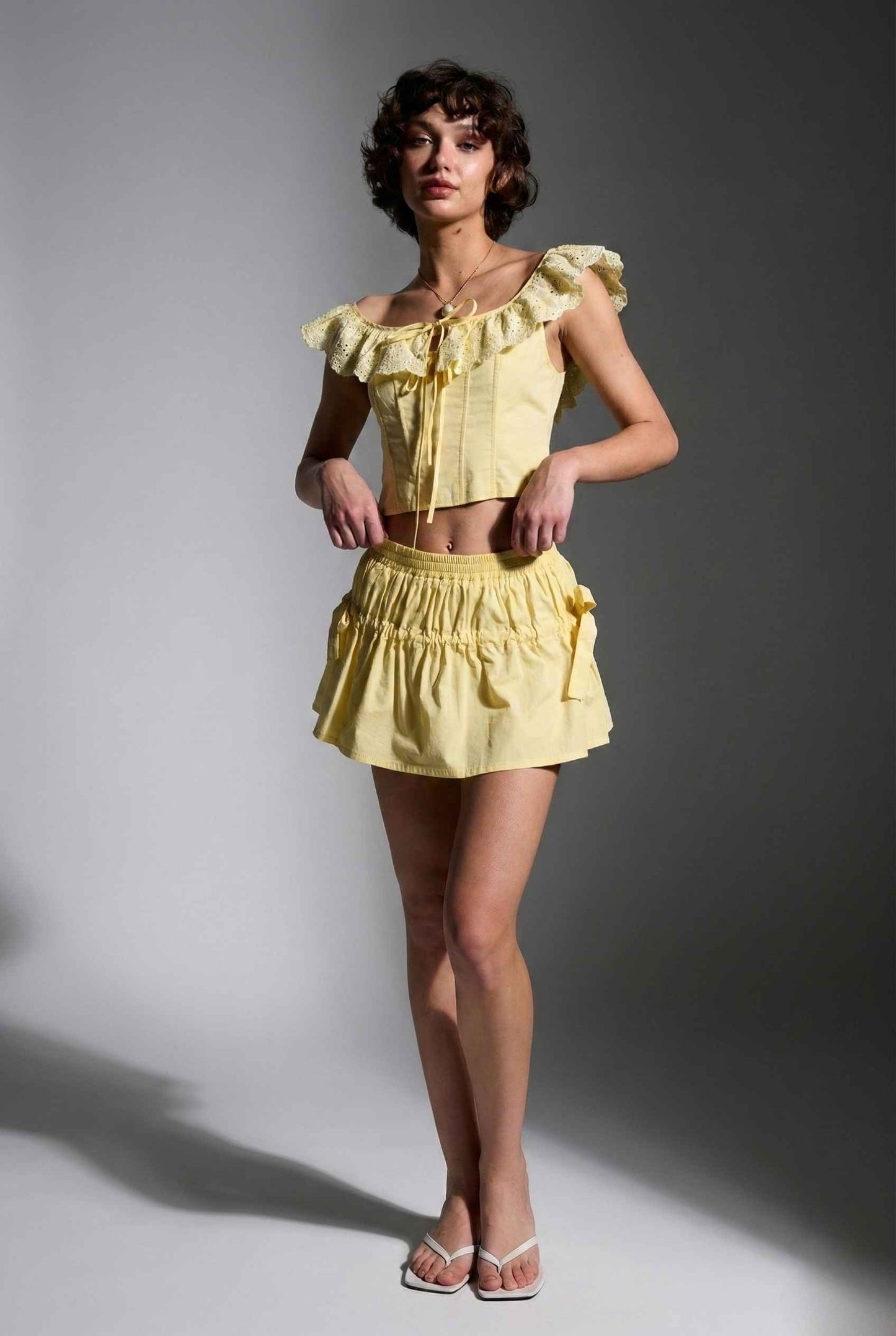 Woman wearing a Clementine Top Lemon Yellow Cotton Eyelet Ruffle Corset and skirt against a gray background
