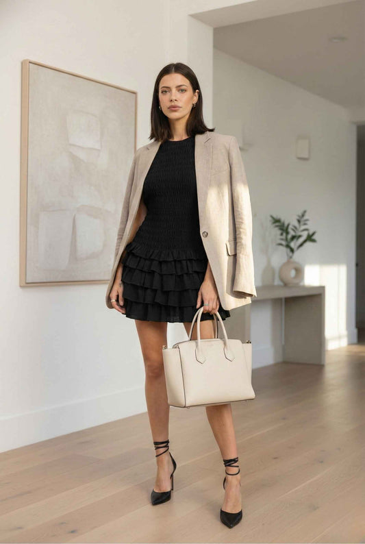 Model wearing Cleo Mini Black Smocked Ruffle Sleeveless Dress With Ruffle in Black with blazer structure tote and heels standing in minimalist luxury home