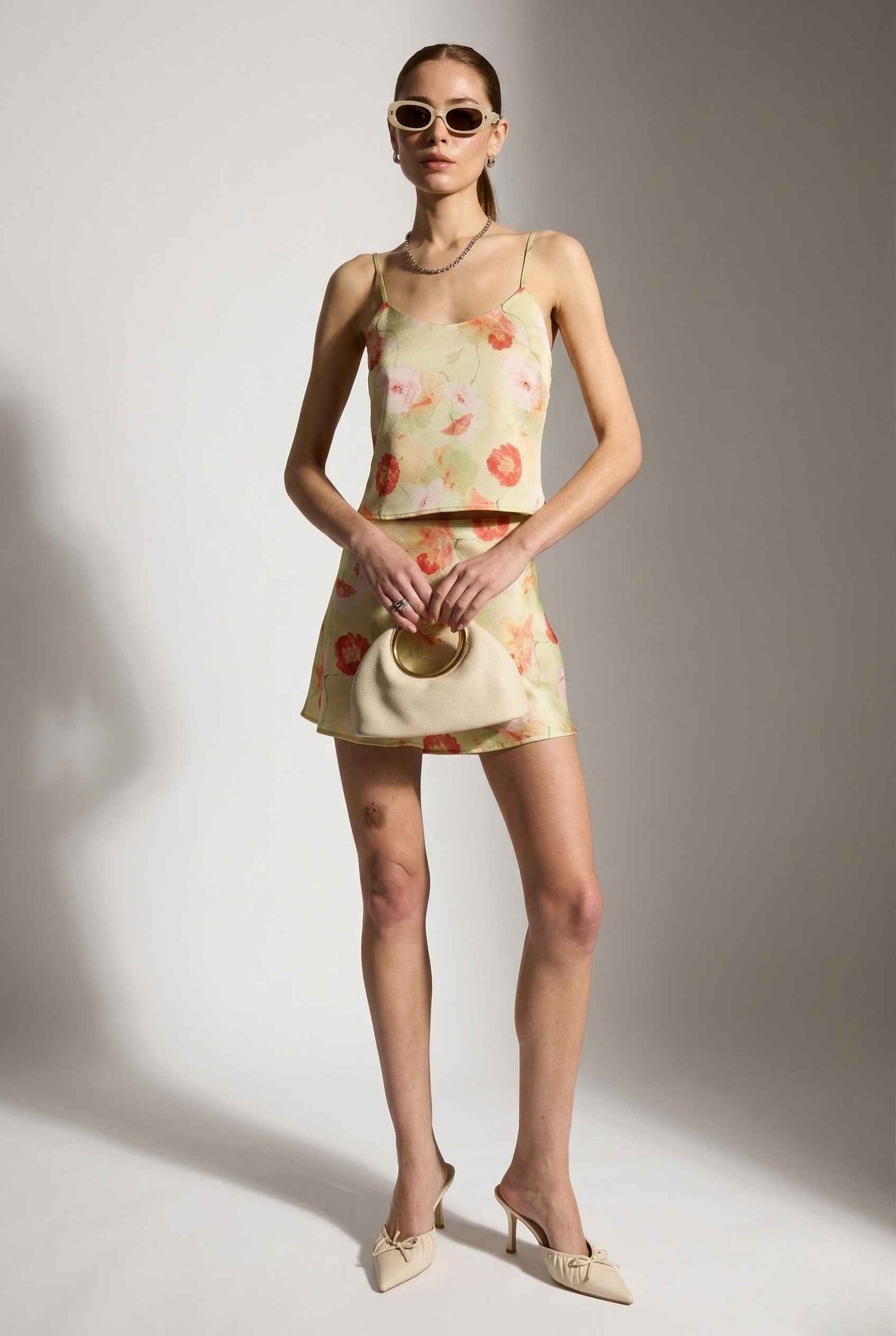 Woman wearing a Cleo Skirt Light Green Floral Bias-Cut Satin Mini Skirt and sunglasses against a plain background