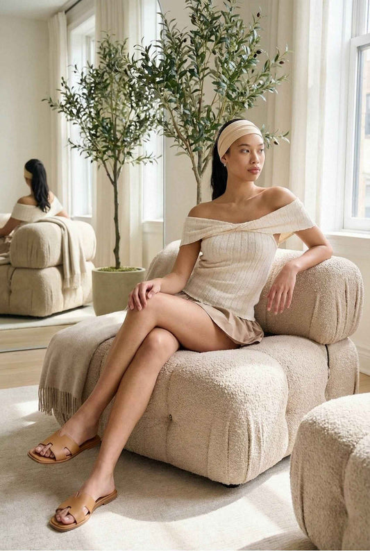 Model wearing Colette Top in Ivory Off-Shoulder Twist Front Short Sleeve Knit Top with ruffle mini skirt and sandals sitting in luxury minimalist living room
