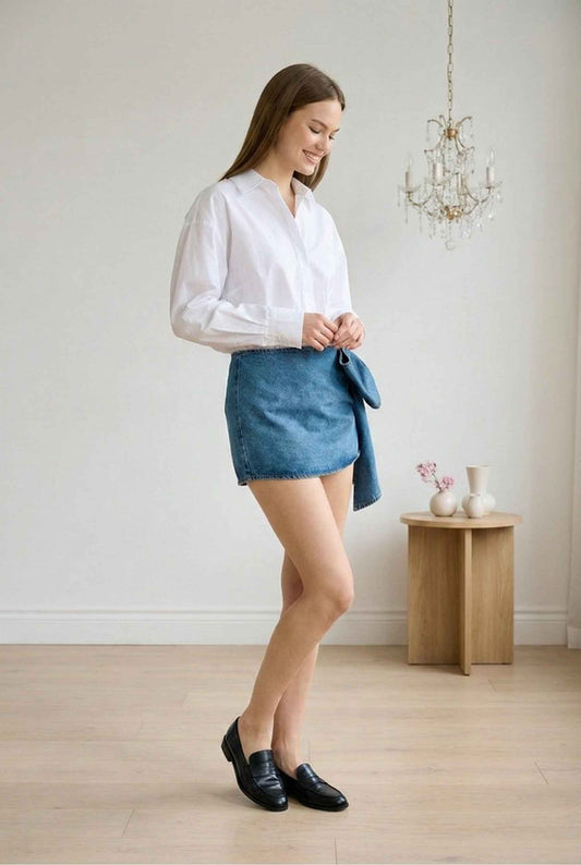 Model wearing Darcy Skirt in Medium Wash Cotton Denim Mini Skirt with Bow paired with tucked in blouse and loafers standing in minimalist room