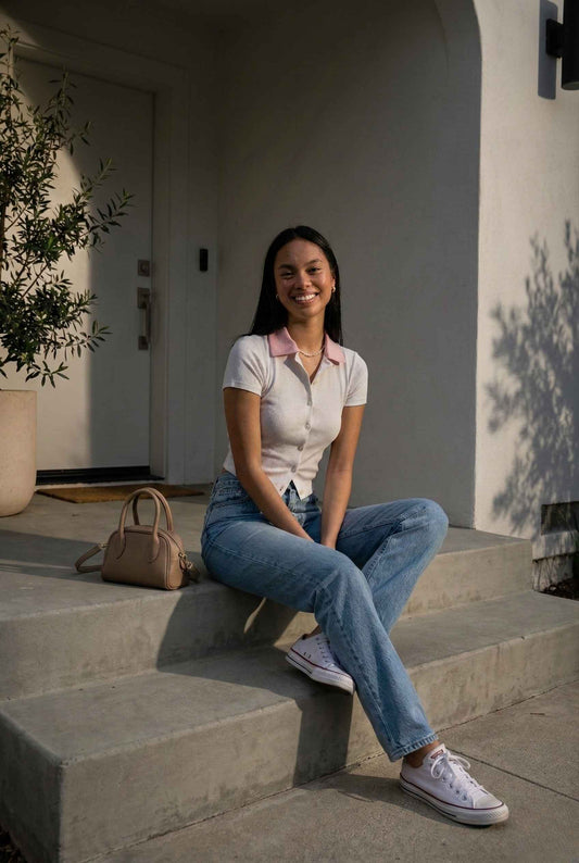 Woman wearing a Darcy Top White & Pink Contrast Collar Button Down Knit sitting on steps outside a building with a plant and handbag in the background