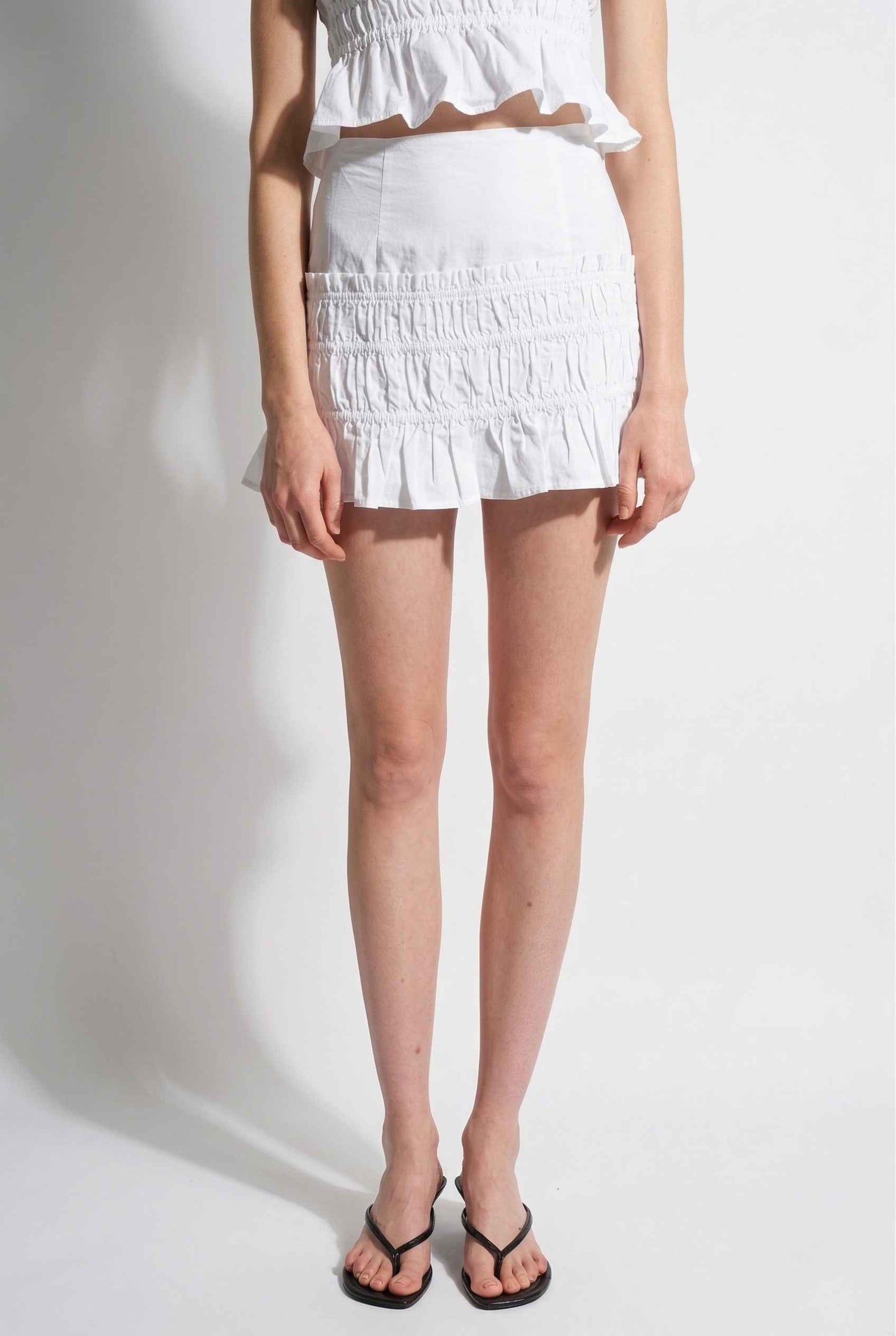 Woman wearing a Elodie Skirt White Cotton Smocked Ruffle Mini Skirt on a plain background