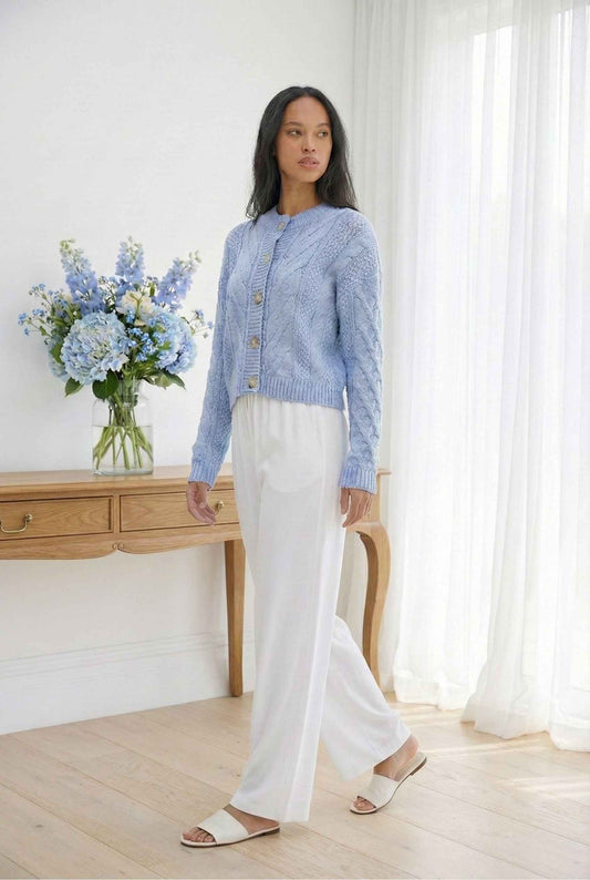 Model wearing Elowen Top in Sky Blue Chunky Cable Knit Button-Front Cardigan with linen pants and slides standing in minimalist room