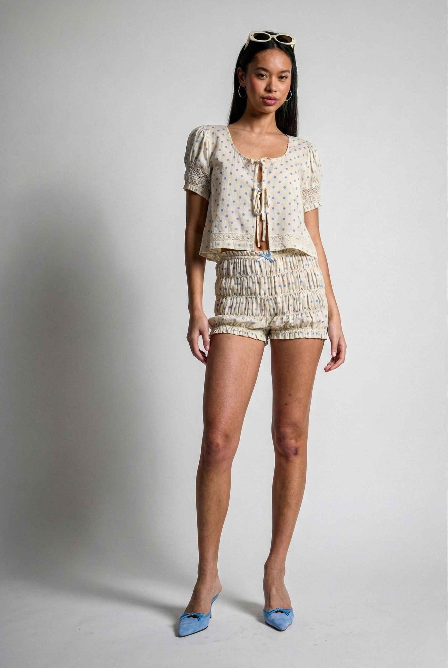 Woman wearing a Elsie Top Ivory & Blue Polka Dot Linen Tie-Front Blouse with shorts and a top on a plain background