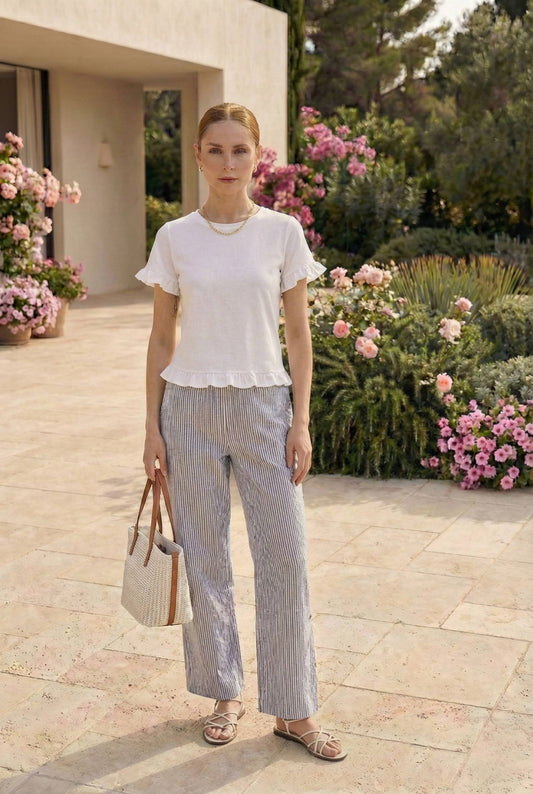 Model wearing Finley Pants in Light Blue Striped Cotton-Blend Trousers with t-shirt and sandals standing in luxury yard with flowers