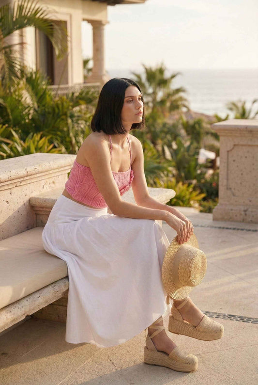 Model wearing Flora Top in Pink Ribbed Knit Halter Crop with 3D Rose Detail paired with flowy maxi skirt and espadrilles sitting on luxury vacation patio by ocean