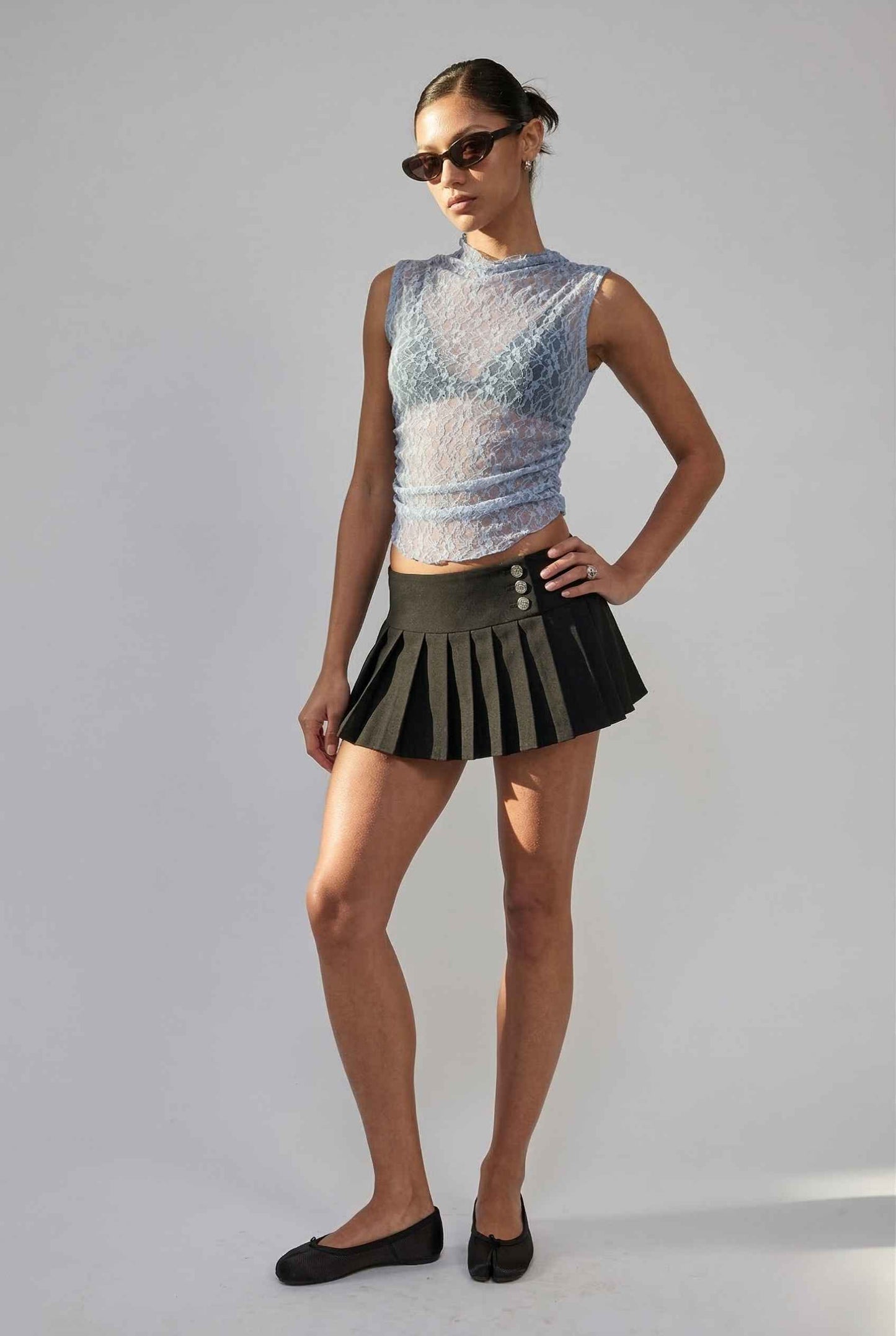 Woman wearing a Iris Top Dusty Blue Sheer Lace Sleeveless Mock Neck and black pleated skirt on a plain background