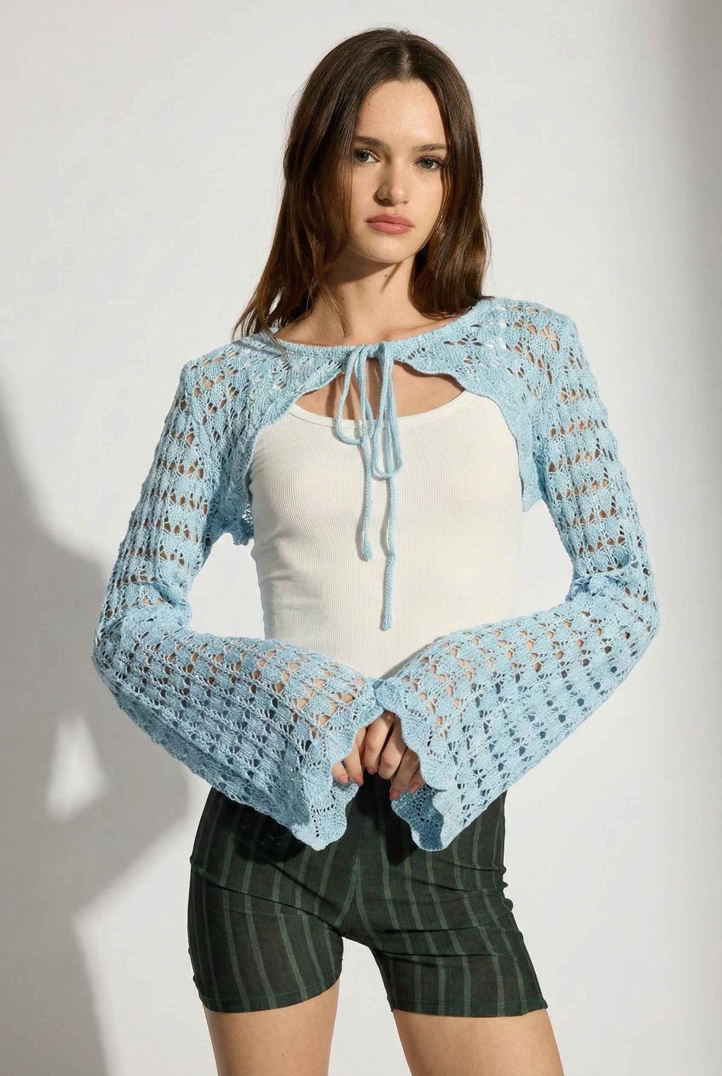 Model wearing Iris Top in Light Blue Crochet Tie-Front Shrug with Bell Sleeves with tank top and striped shorts on a plain background