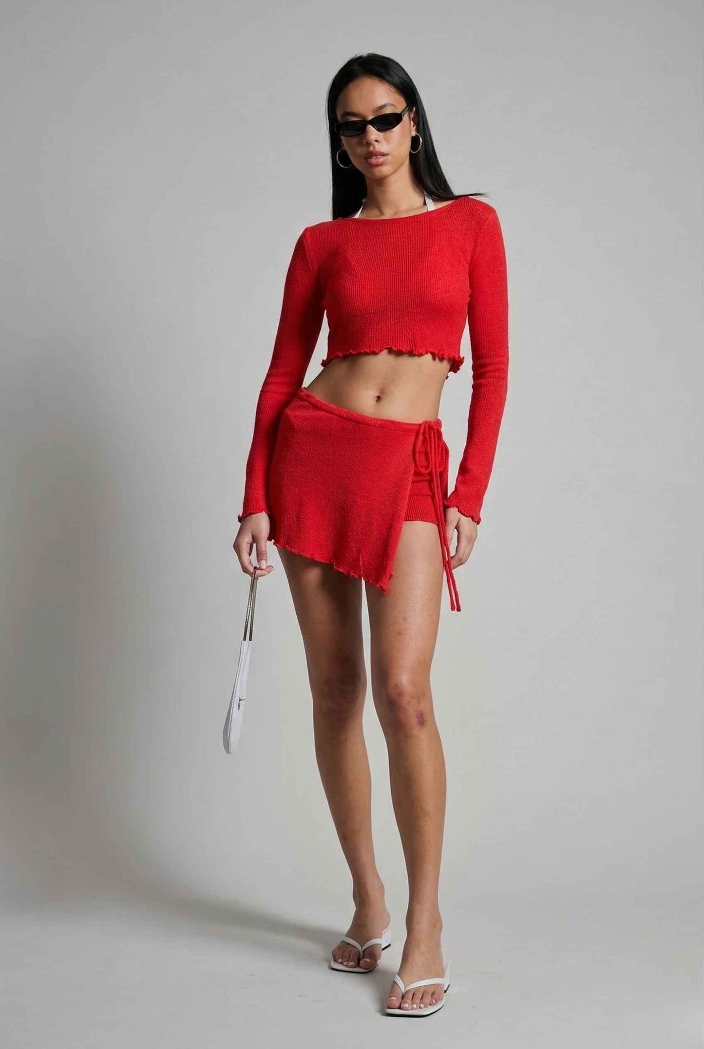 Woman wearing a Kaia Top Red Sheer Knit Boat Neck Long Sleeve Crop with sunglasses on a gray background