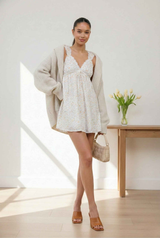 Woman in a Liana Mini Cream Floral Lace Babydoll Dress and cardigan standing in a bright room with a vase of flowers on a table.