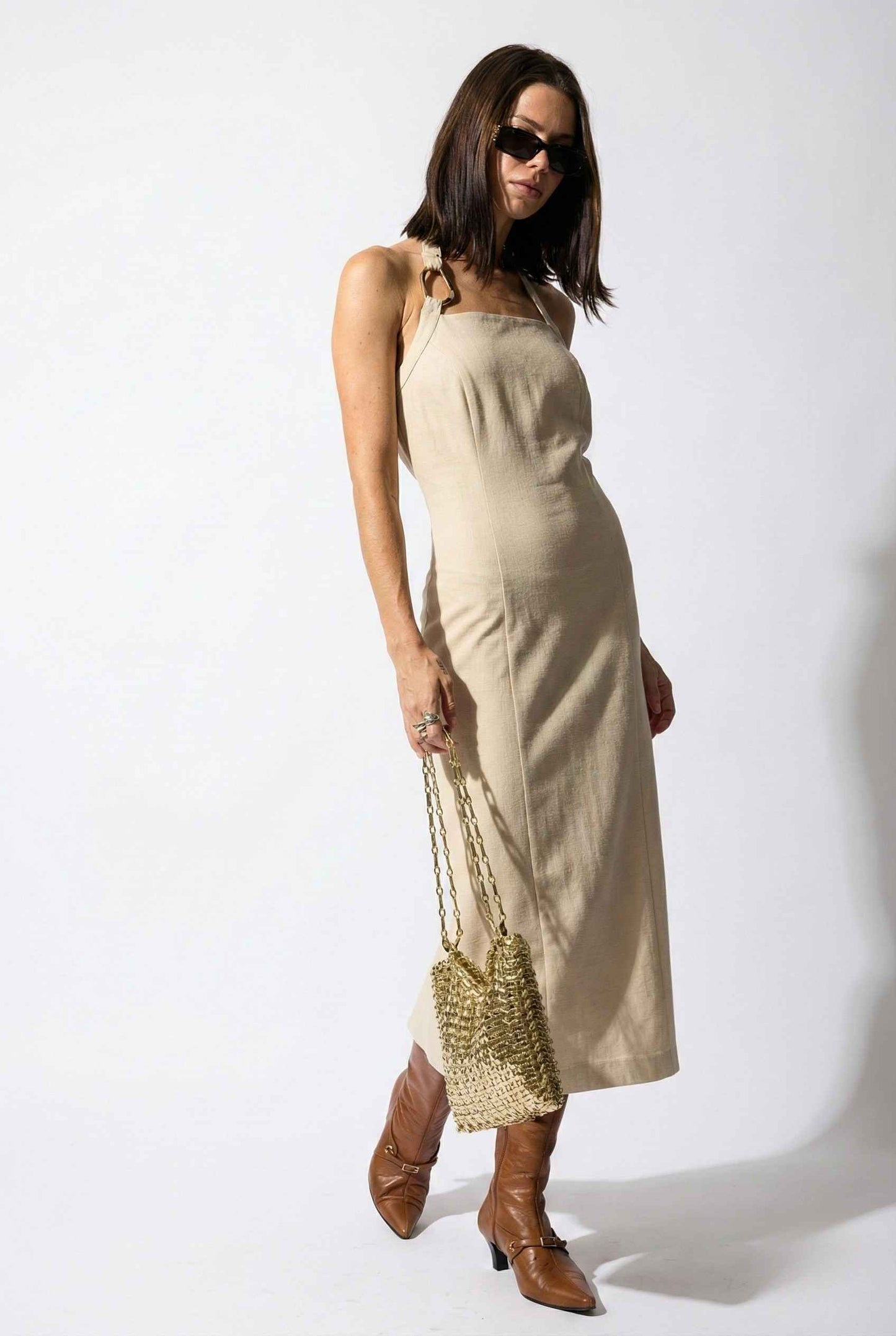 Woman wearing a Mercer Midi Taupe Halter Hardware Dress with sunglasses and holding a woven bag on a white background