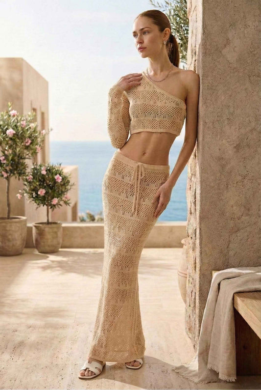 Model wearing Nola Top in Cream Cotton-Blend Crochet One-Shoulder Long Sleeve with matching maxi skirt and sandals standing on luxury vacation balcony by ocean