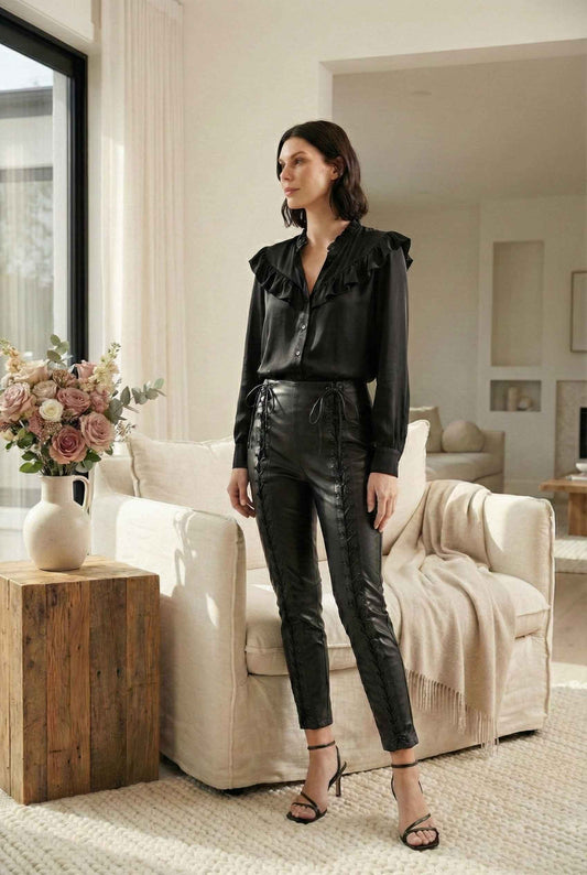 Model wearing Nova Pants in Black Vegan Leather High-Waist Lace-Up Trousers with ruffle blouse and strappy sandals standing in luxury living room