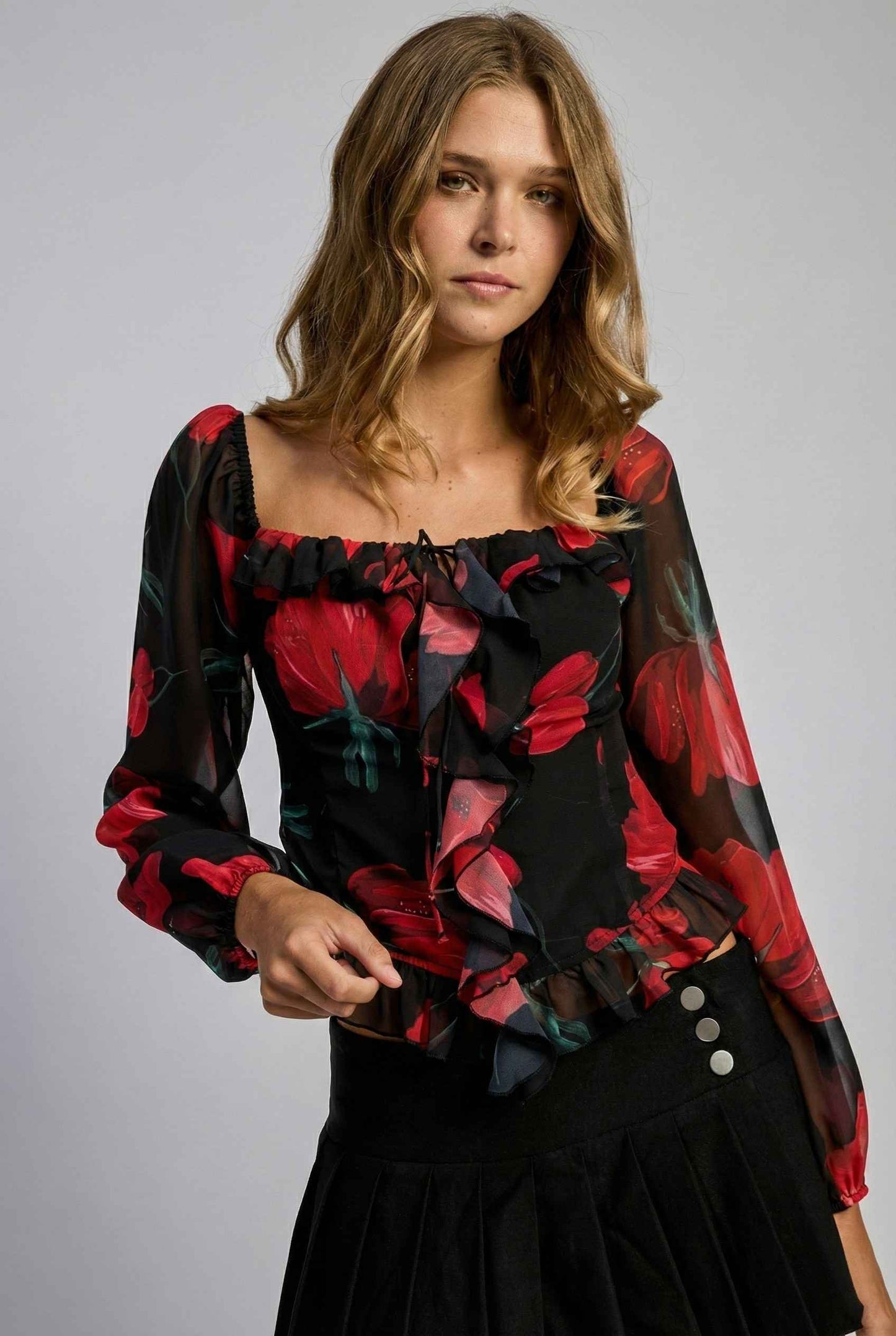 Woman wearing a Rosalia Blouse Black & Red Rose Print Ruffle Top with red floral patterns on a gray background