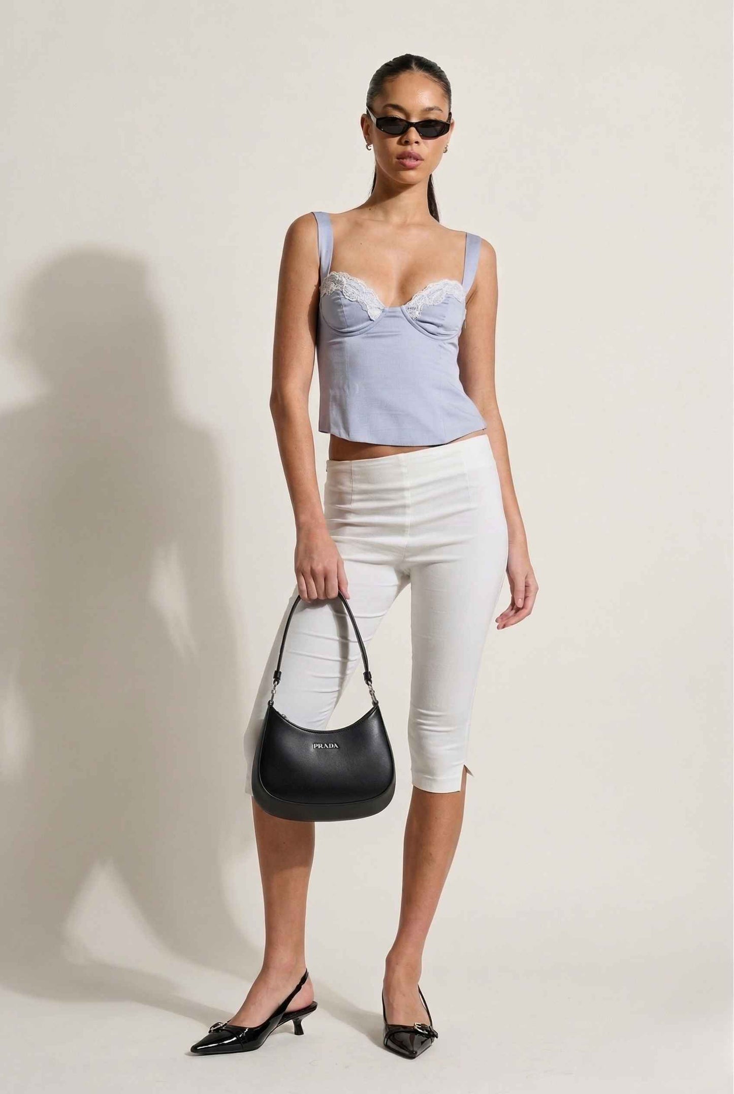 Woman wearing a Sophie Top Blue Pastel Satin Corset Cami with White Lace Trim and white pants, holding a black handbag against a plain background