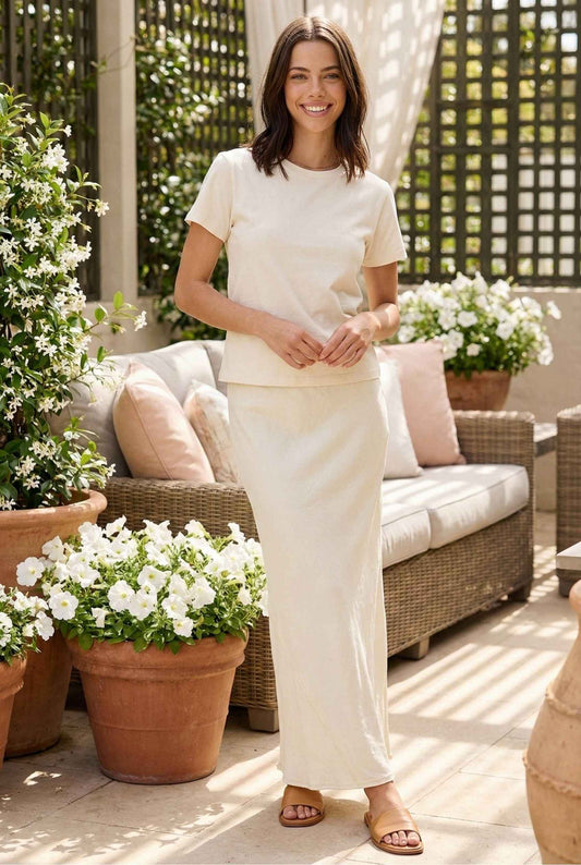 Model wearing Veda Skirt in Cream Satin Drawstring Maxi Skirt paired with basic t-shirt and leather slides posing on patio