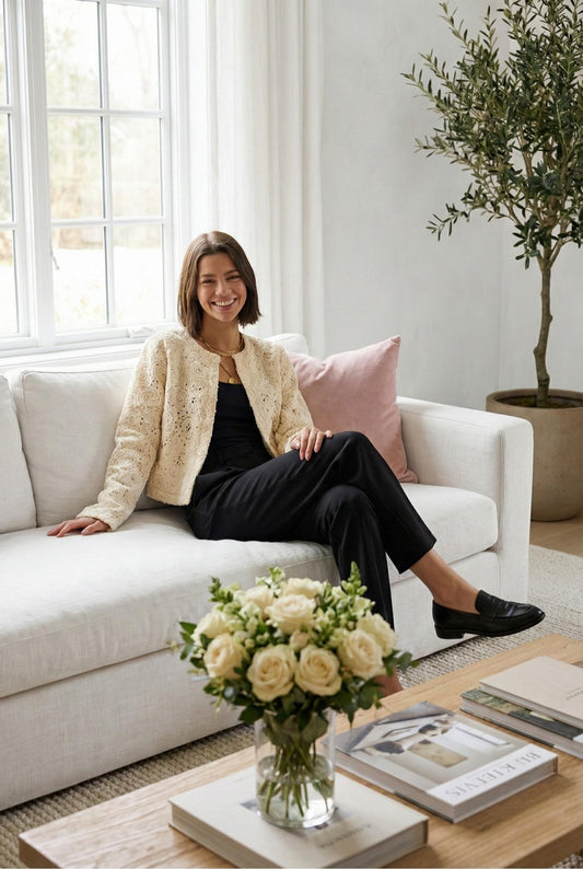 Model wearing Emory Park Macrame Cropped Jacket in cream with intricate floral knit design sitting on couch in luxury living room roses