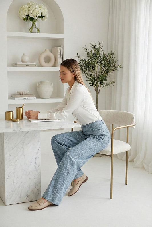 Model wearing Emory Park Mid Rise Wide Leg Pants in blue Denim pinstripe with blouse and loafers sitting in luxury minimalist office