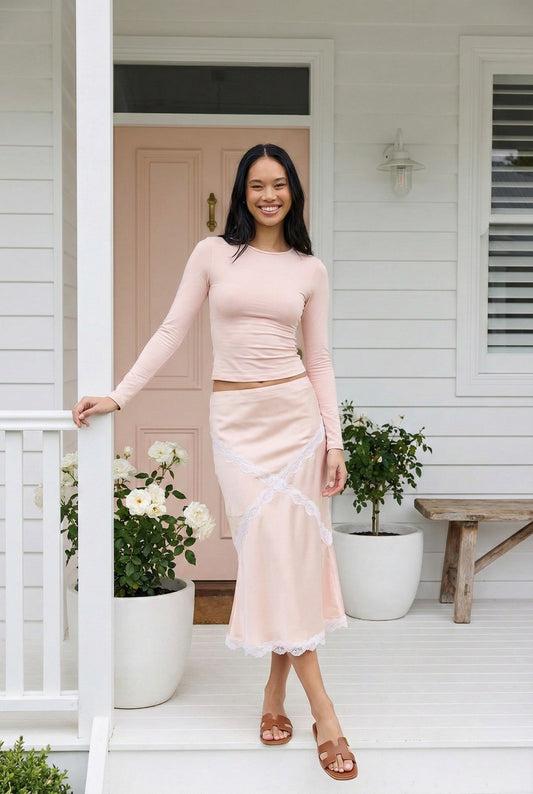 Model wearing Emory Park Midi Satin Skirt With Lace Detail in blush pink standing outside feminine white luxury home