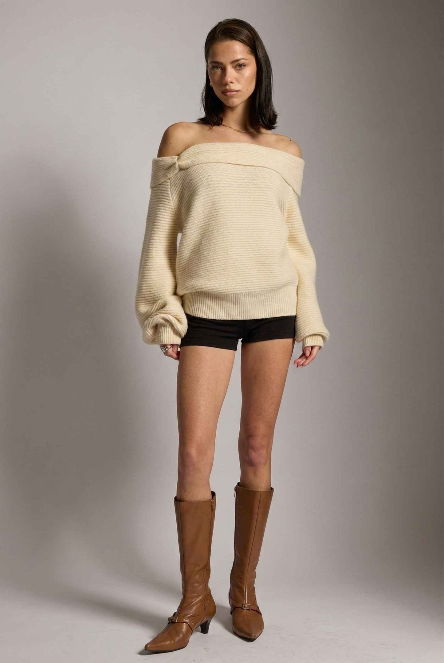 Model wearing Amara Top in Cream Ribbed Knit Twist-Front Off-Shoulder Sweater with micro shorts and tall boots