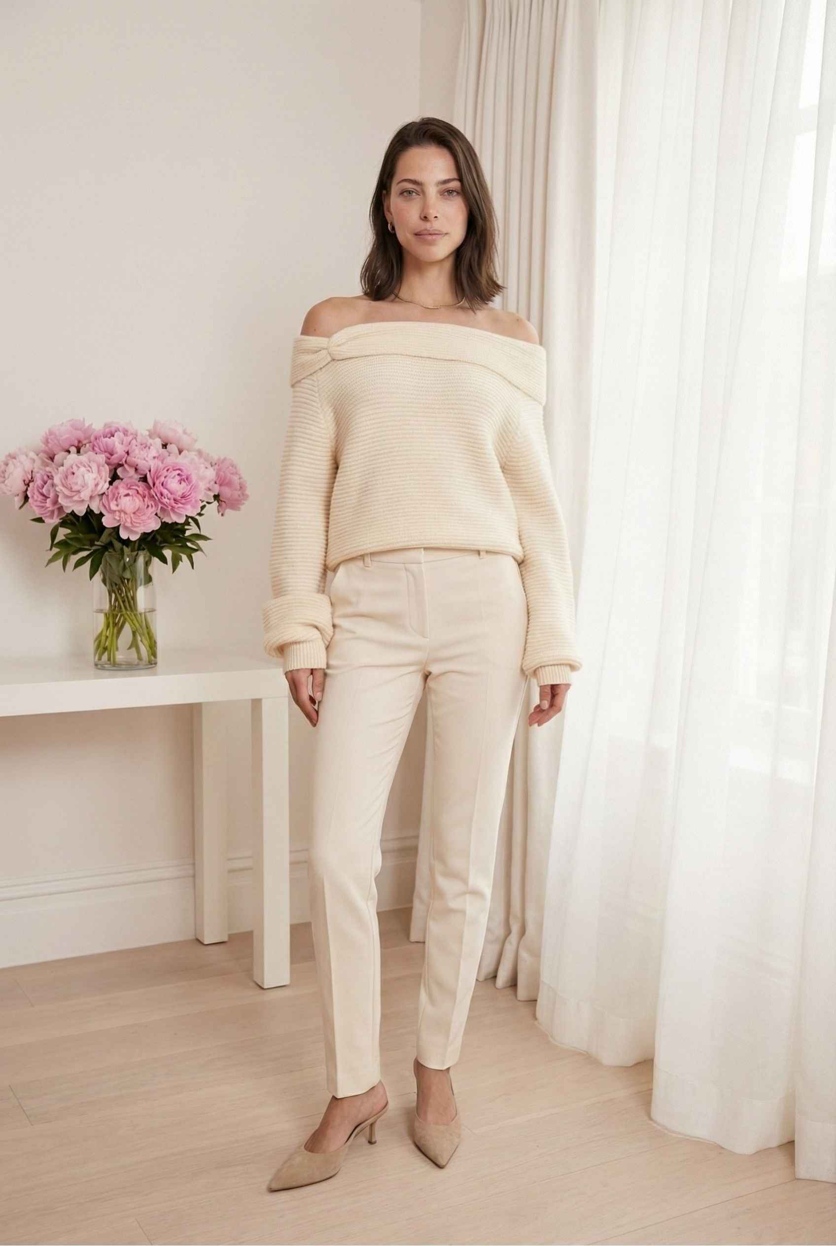Model wearing Amara Top in Cream Ribbed Knit Twist-Front Off-Shoulder Sweater with trousers and pointed heels standing in feminine minimalist room