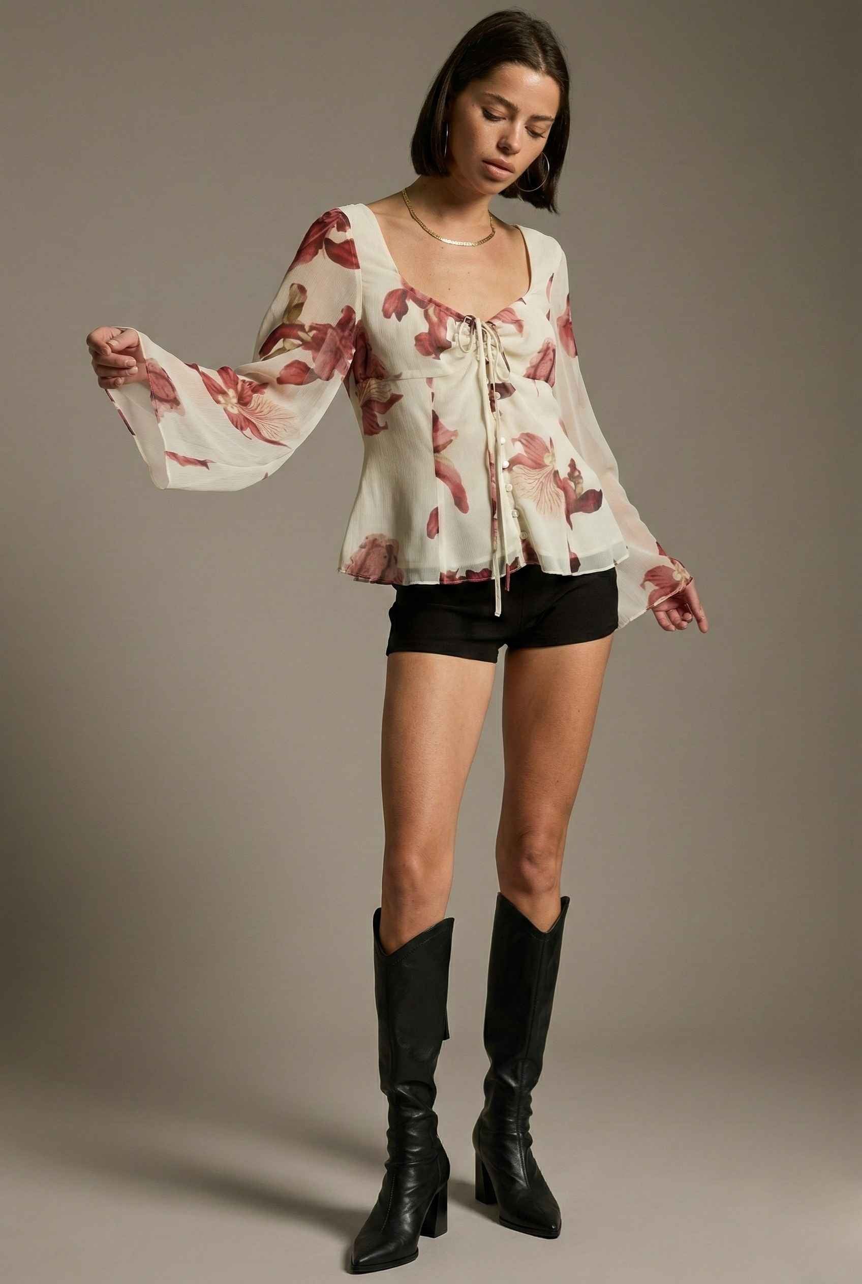Woman wearing a Aria Top Ivory Floral Tie-Front Bell Sleeve Blouse, black shorts, and black knee-high boots against a plain background