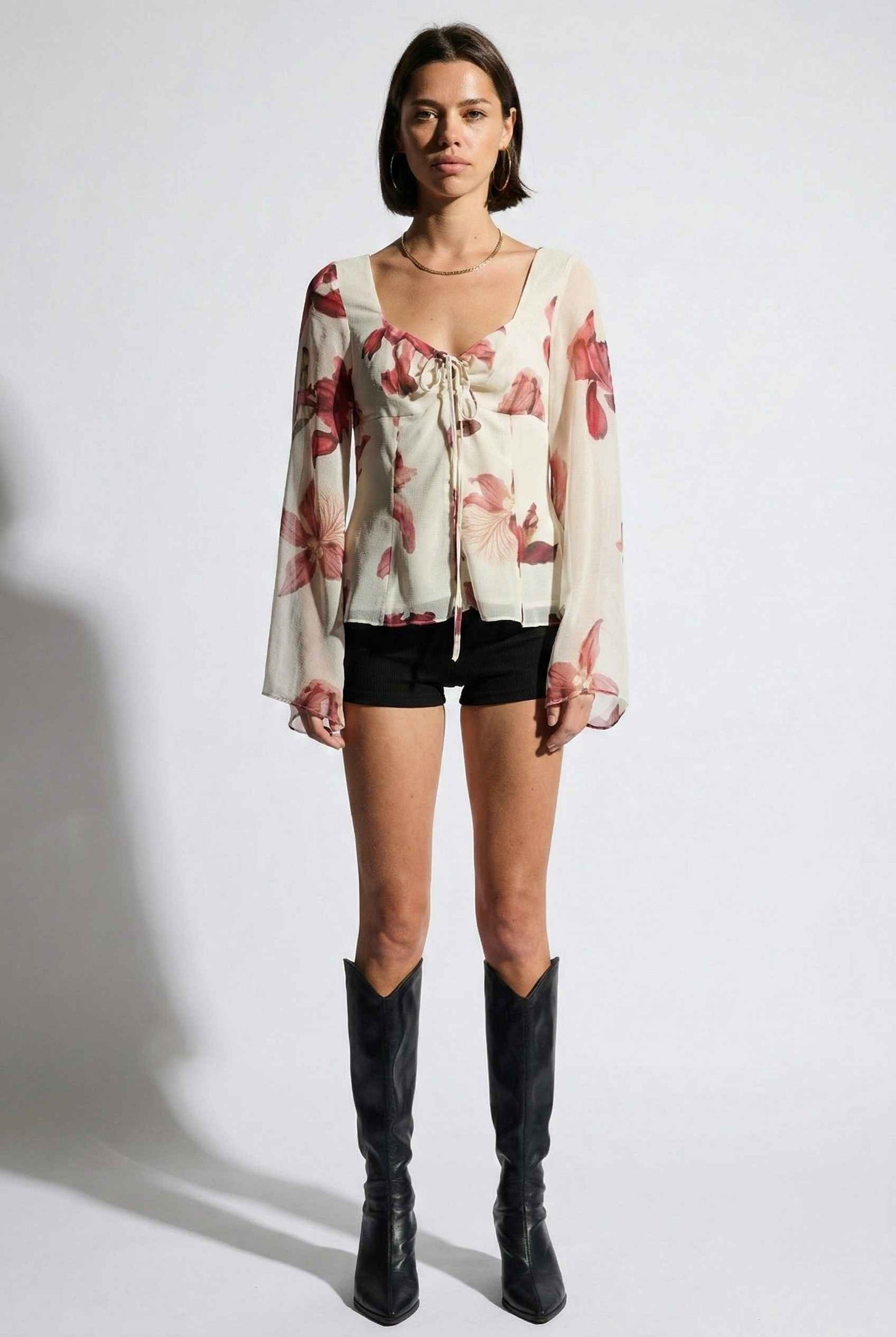 Woman wearing a Aria Top Ivory Floral Tie-Front Bell Sleeve Blouse, black shorts, and black knee-high boots on a white background