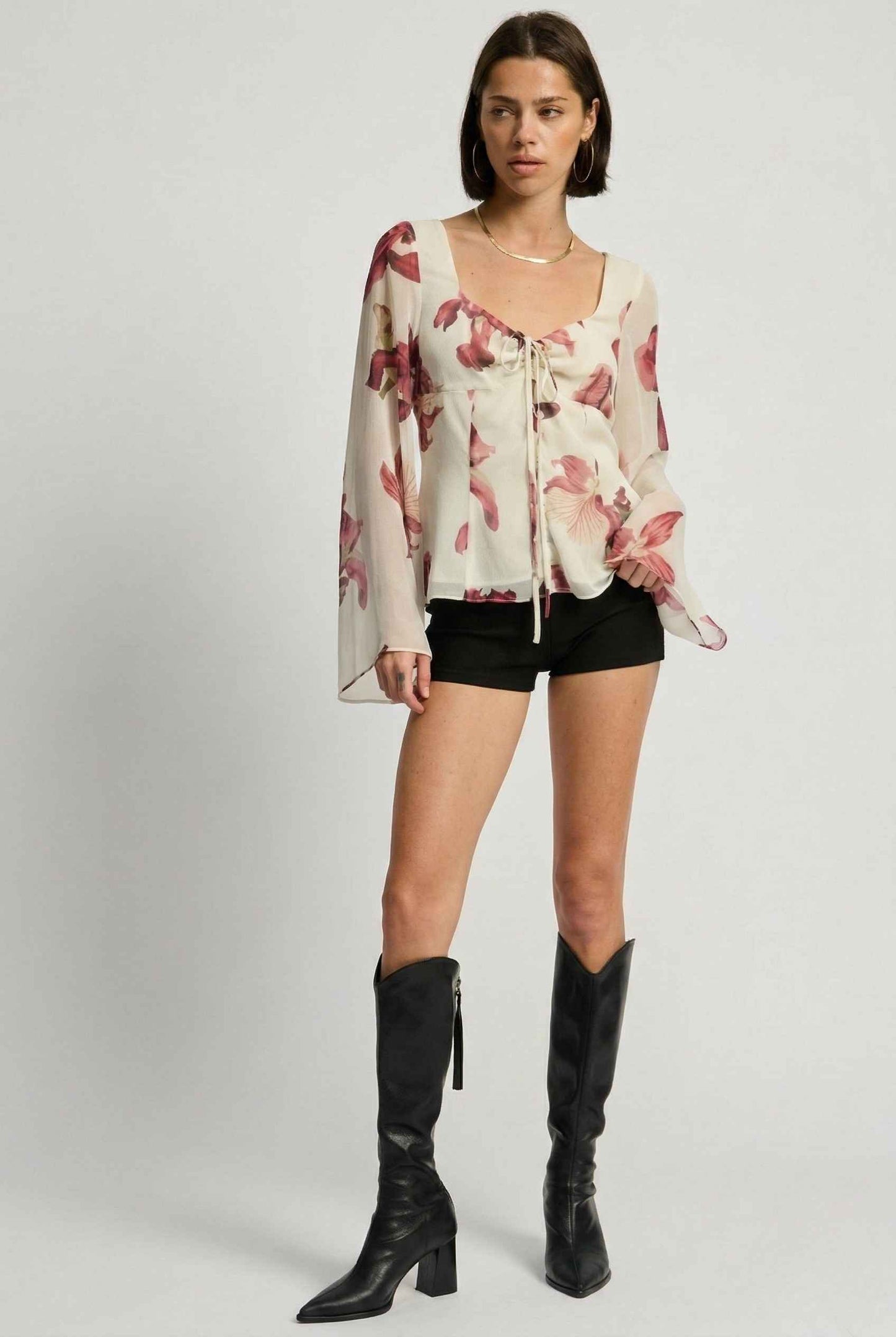 Woman wearing a Aria Top Ivory Floral Tie-Front Bell Sleeve Blouse, black shorts, and knee-high boots on a plain background