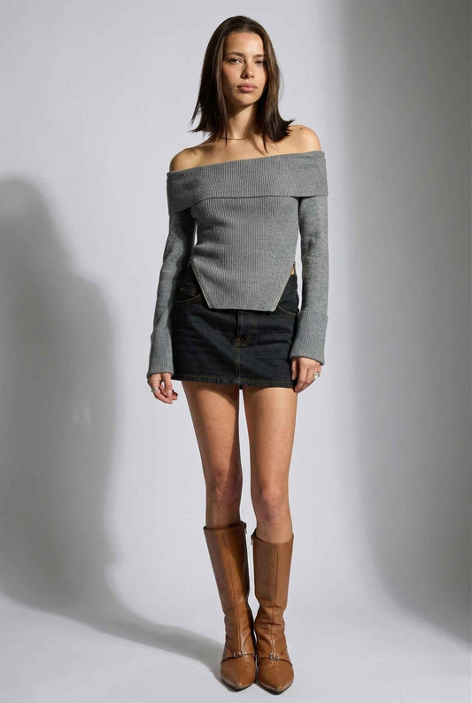 Model wearing Aria Top Ribbed Knit Off-Shoulder Sweater with Side Zipper in Grey paired with denim mini skirt and tall boots on a plain background