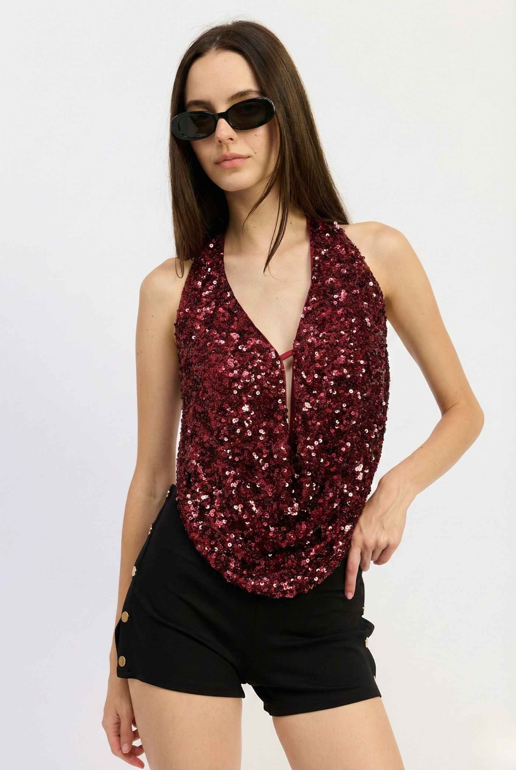 Woman wearing a Astra Top Cherry Sequin Draped Halter Neck and black shorts on a white background