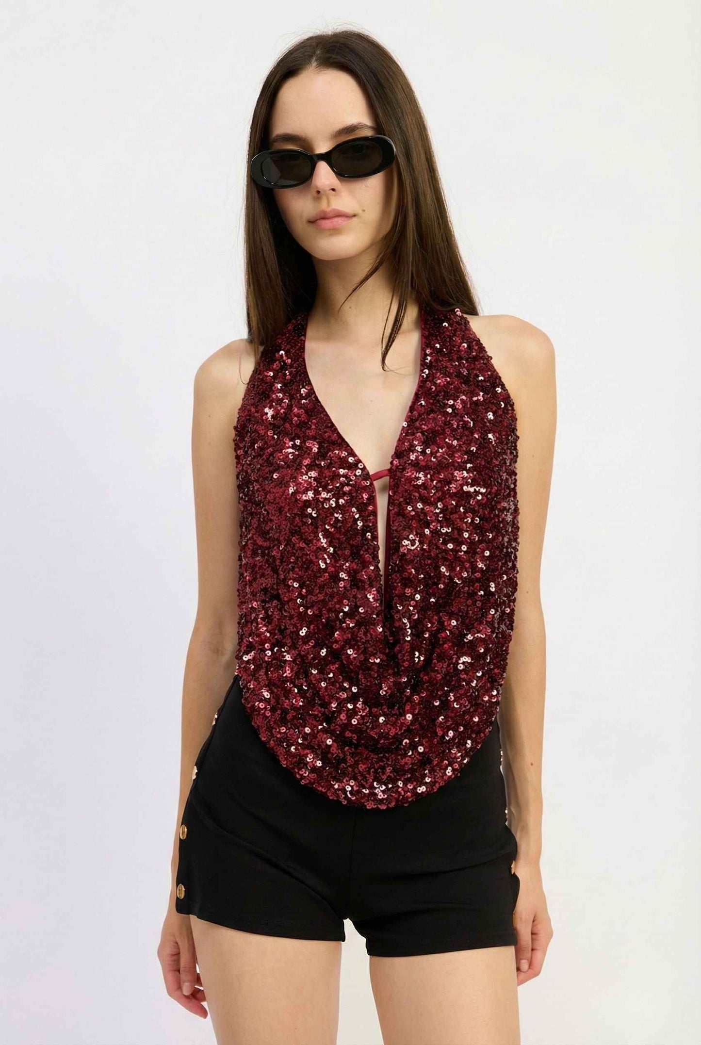 Woman wearing a Astra Top Cherry Sequin Draped Halter Neck and black shorts on a white background