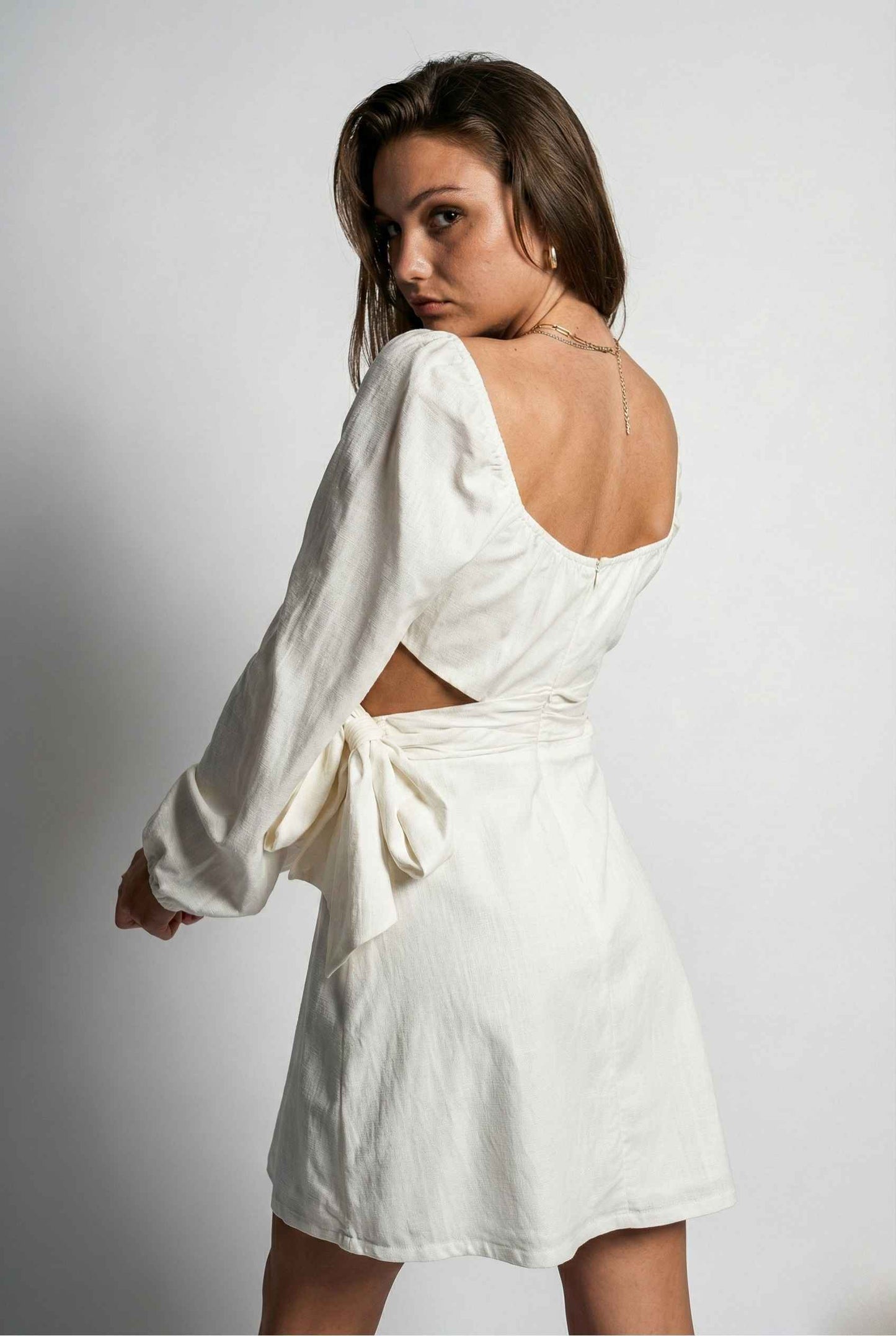 Woman wearing a Aurelia Mini White Long Sleeve Cutout Dress with long sleeves and open back on a plain background