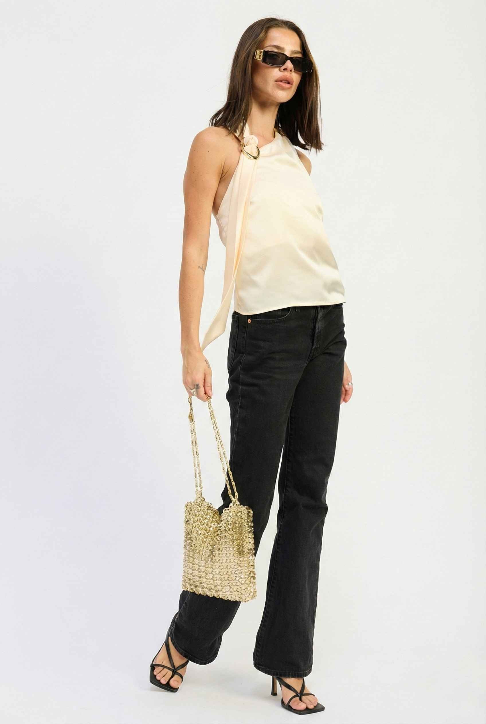 Woman wearing a Aurelia Top Ivory Satin One-Shoulder Scarf Blouse and black pants, holding a straw bag on a white background