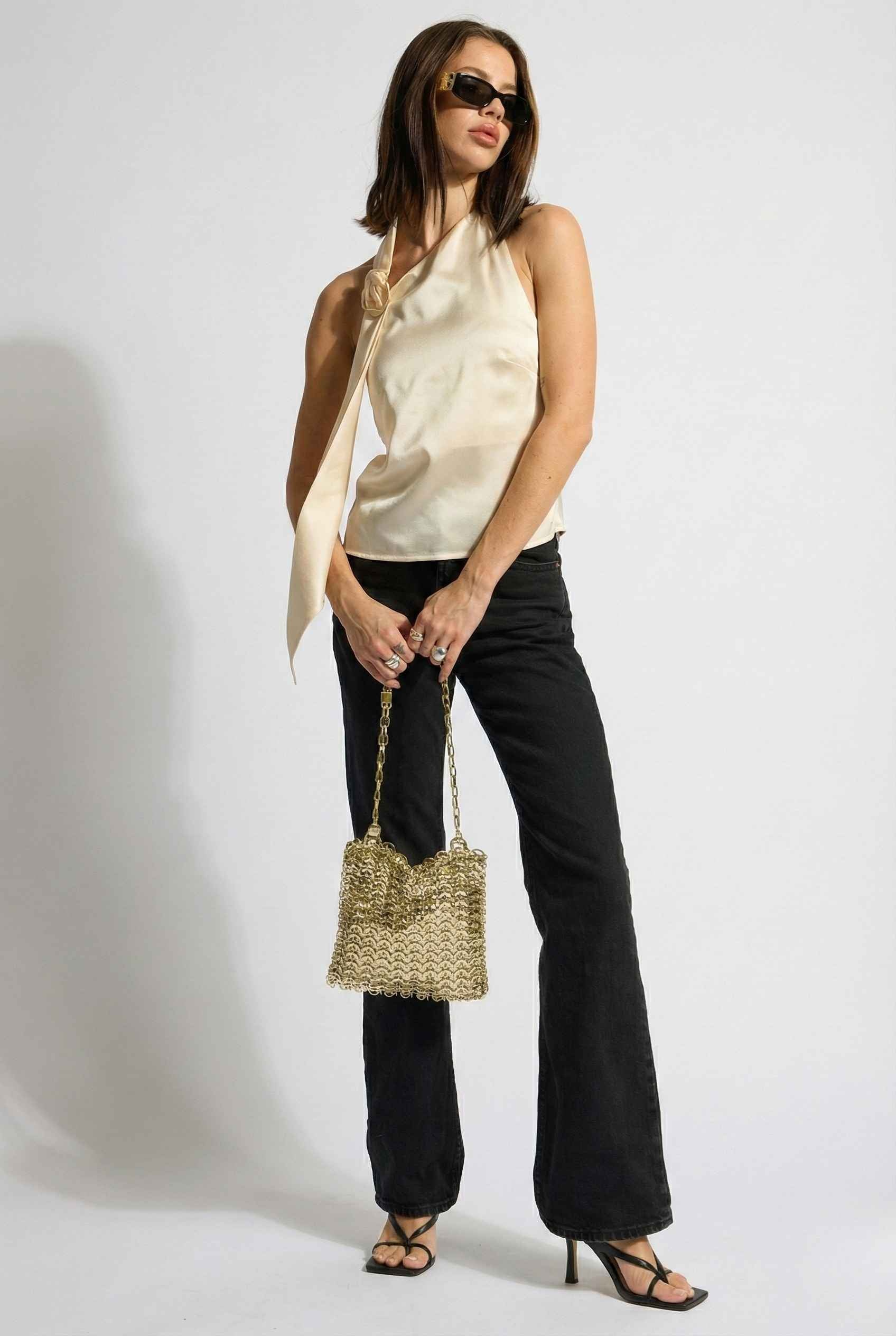 Woman wearing a Aurelia Top Ivory Satin One-Shoulder Scarf Blouse and black pants, holding a straw handbag against a white background