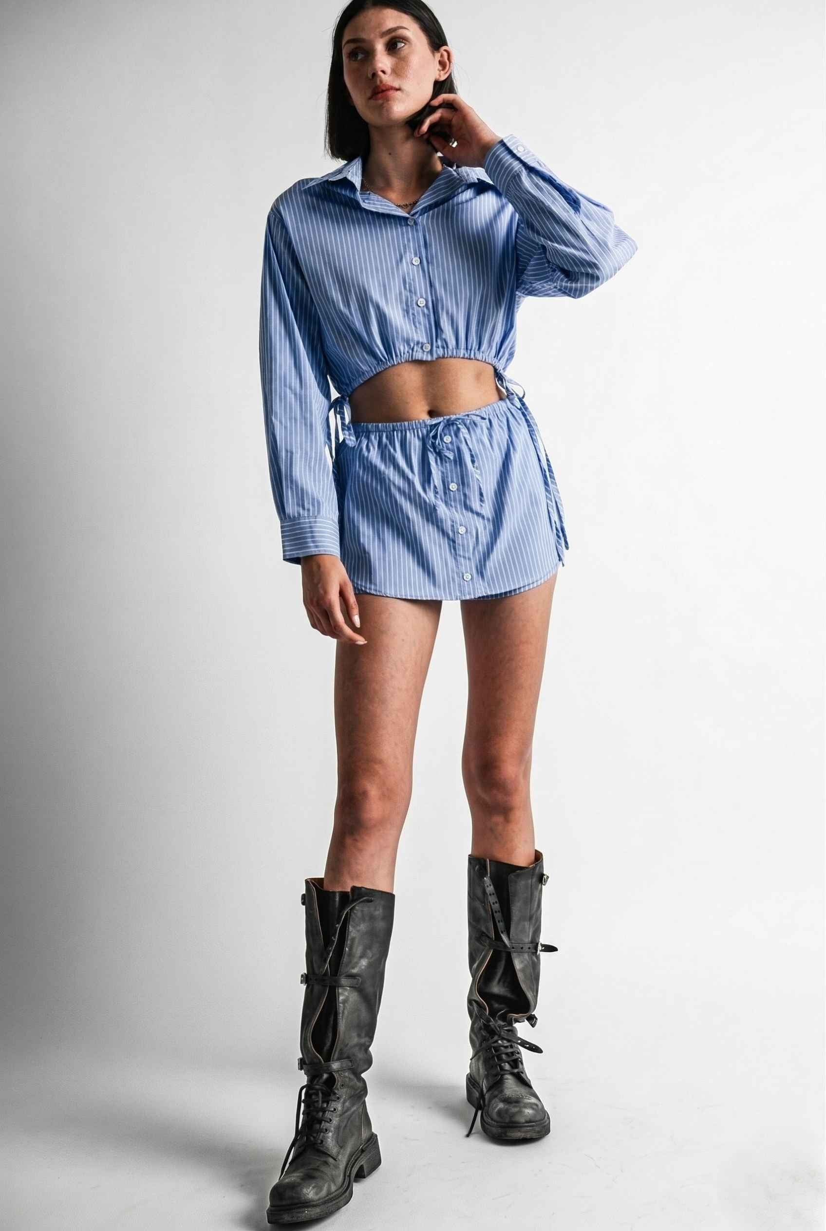 Woman wearing a Blair Skort Blue Striped Poplin Mini Skort with Shirttail Hem with black boots on a plain background