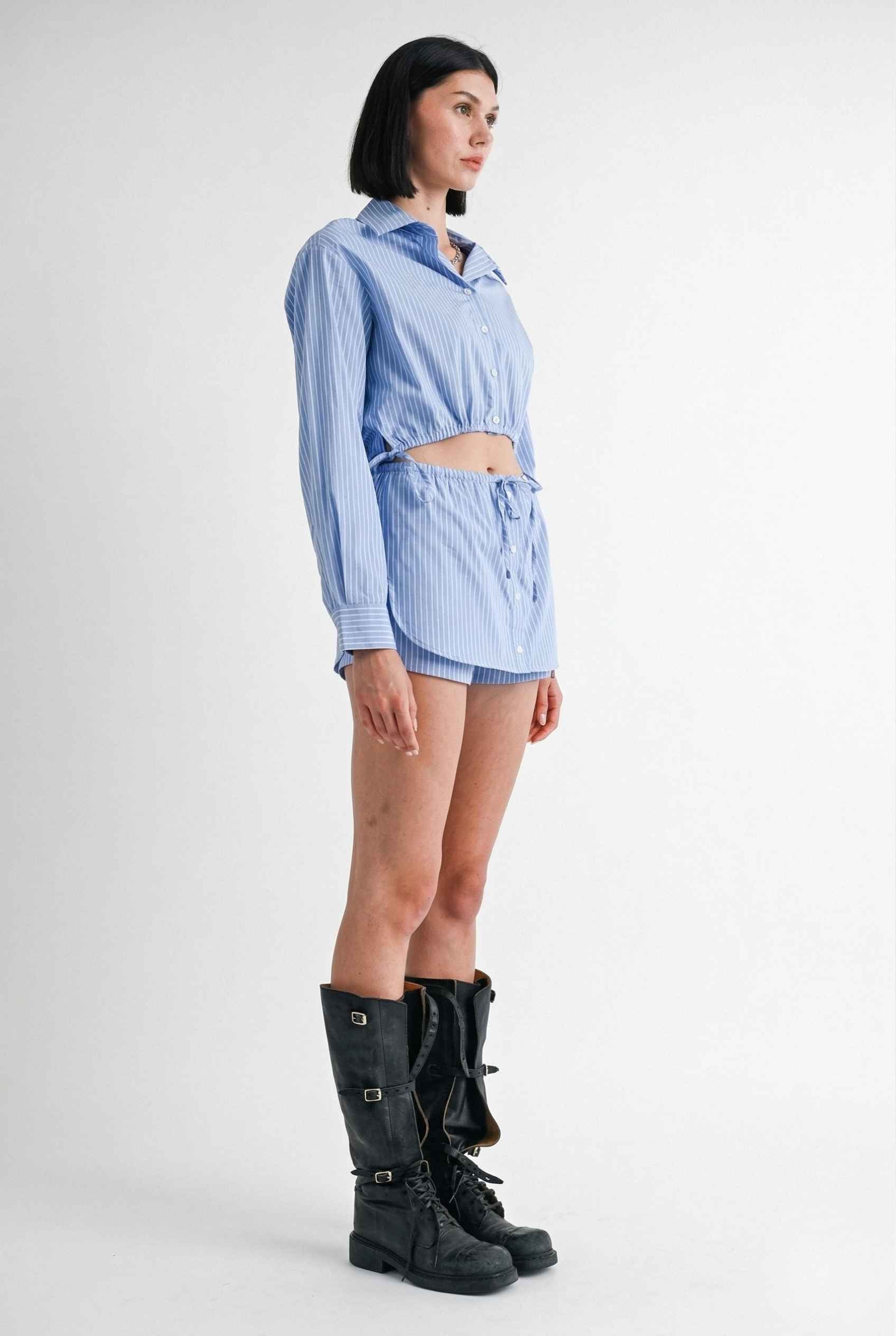 Woman wearing a Blair Skort Blue Striped Poplin Mini Skort with Shirttail Hem with black boots on a plain background
