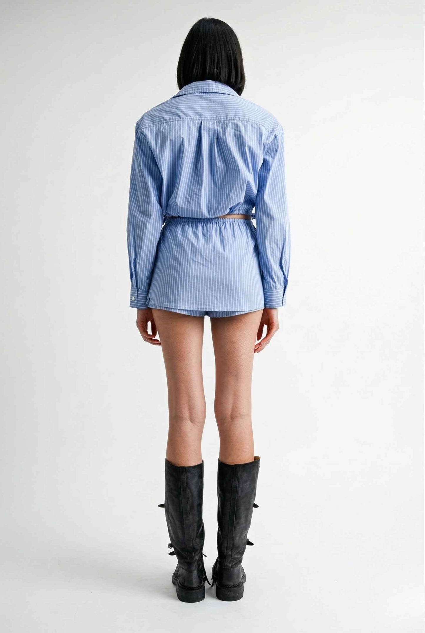 Woman wearing a Blair Skort Blue Striped Poplin Mini Skort with Shirttail Hem with black boots on a plain background