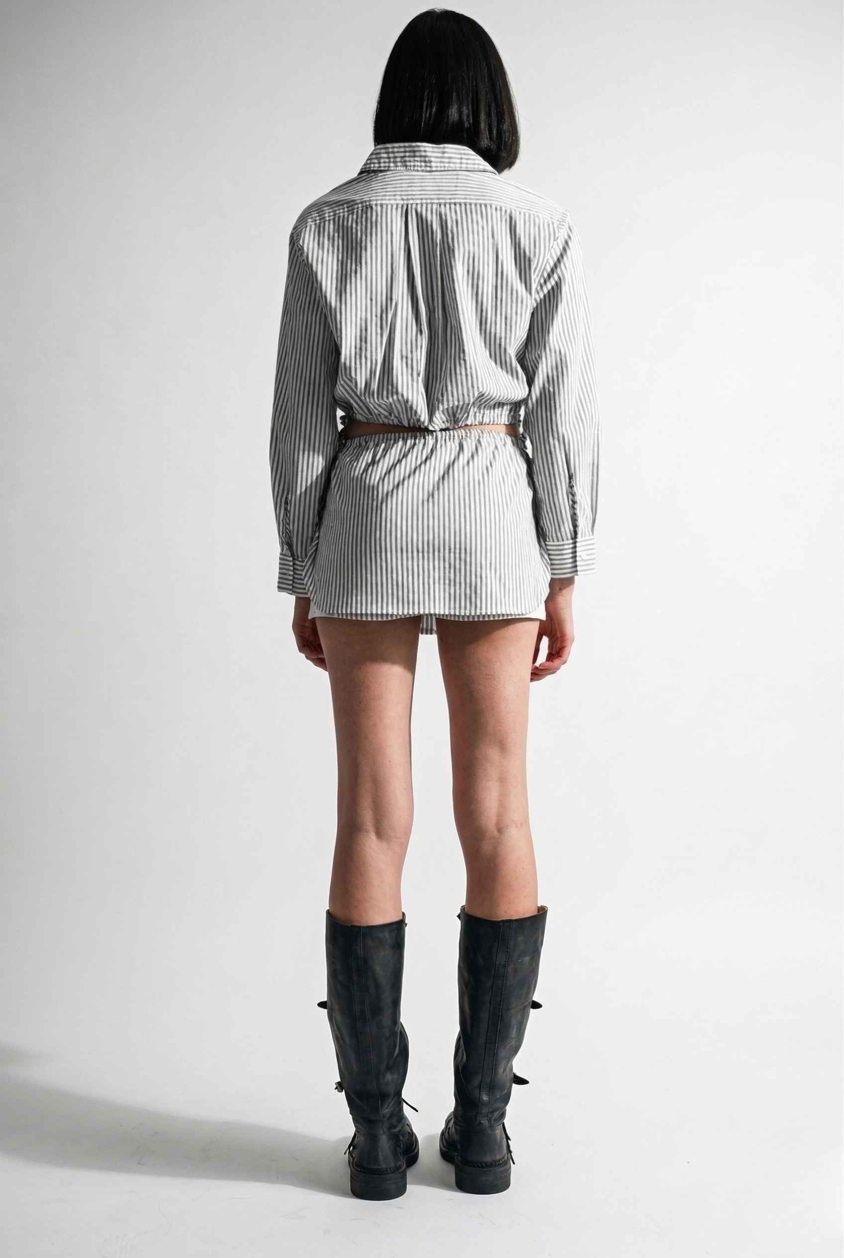 Woman wearing a checkered shirt, Blair Skort White Striped Poplin Mini Skort with Shirttail Hem, and black boots on a white background