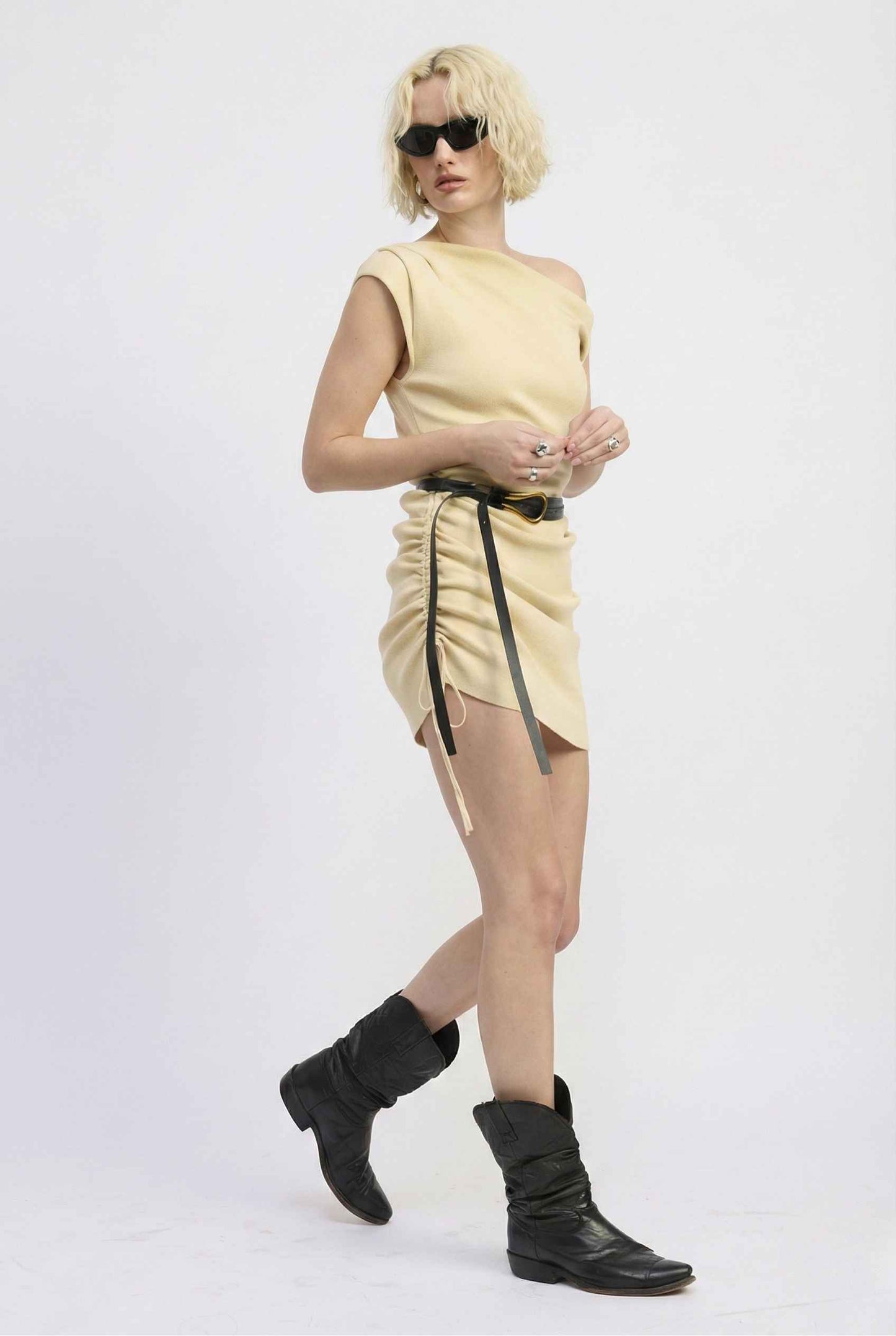 Woman wearing a Blaire Mini Cream Off-Shoulder Ruched Dress with a black belt and black boots on a white background