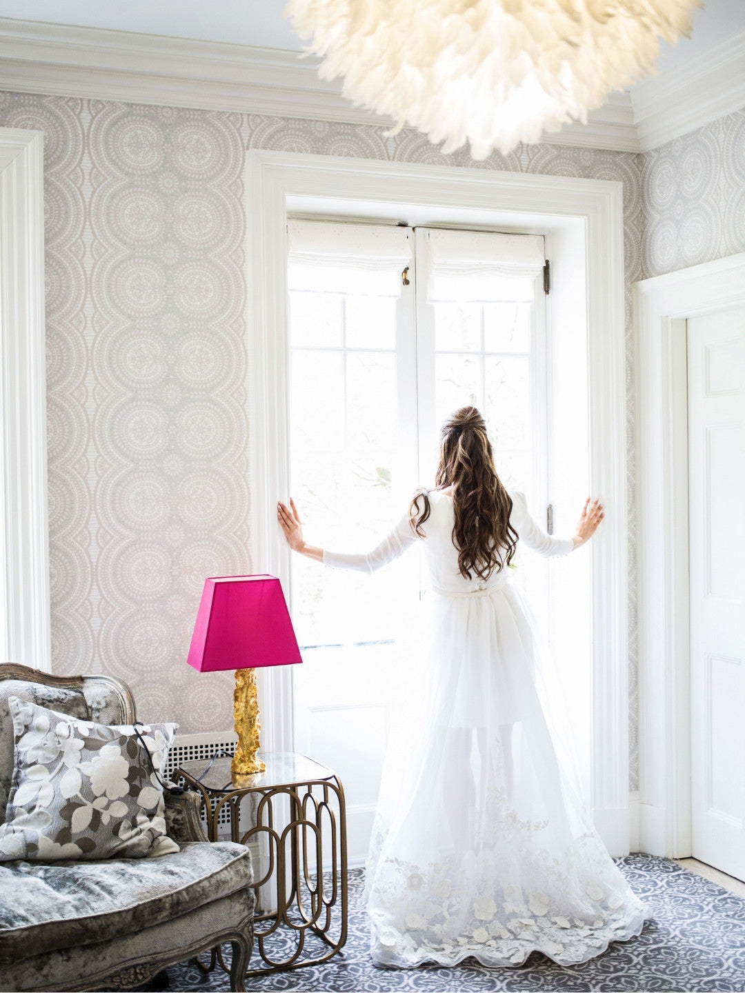 Woman in a white dress standing in a stylish room with patterned wallpaper and a pink lamp.