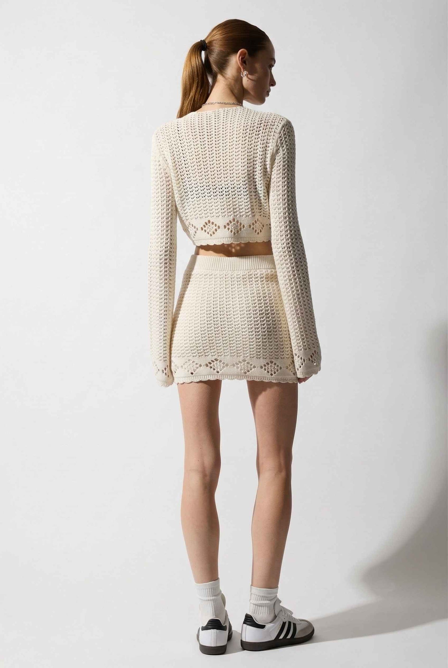 Model wearing Calla Skirt in Ivory Cotton Crochet Mini Skirt with Lettuce Hem paired with matching bolero top and sneakers on a plain background