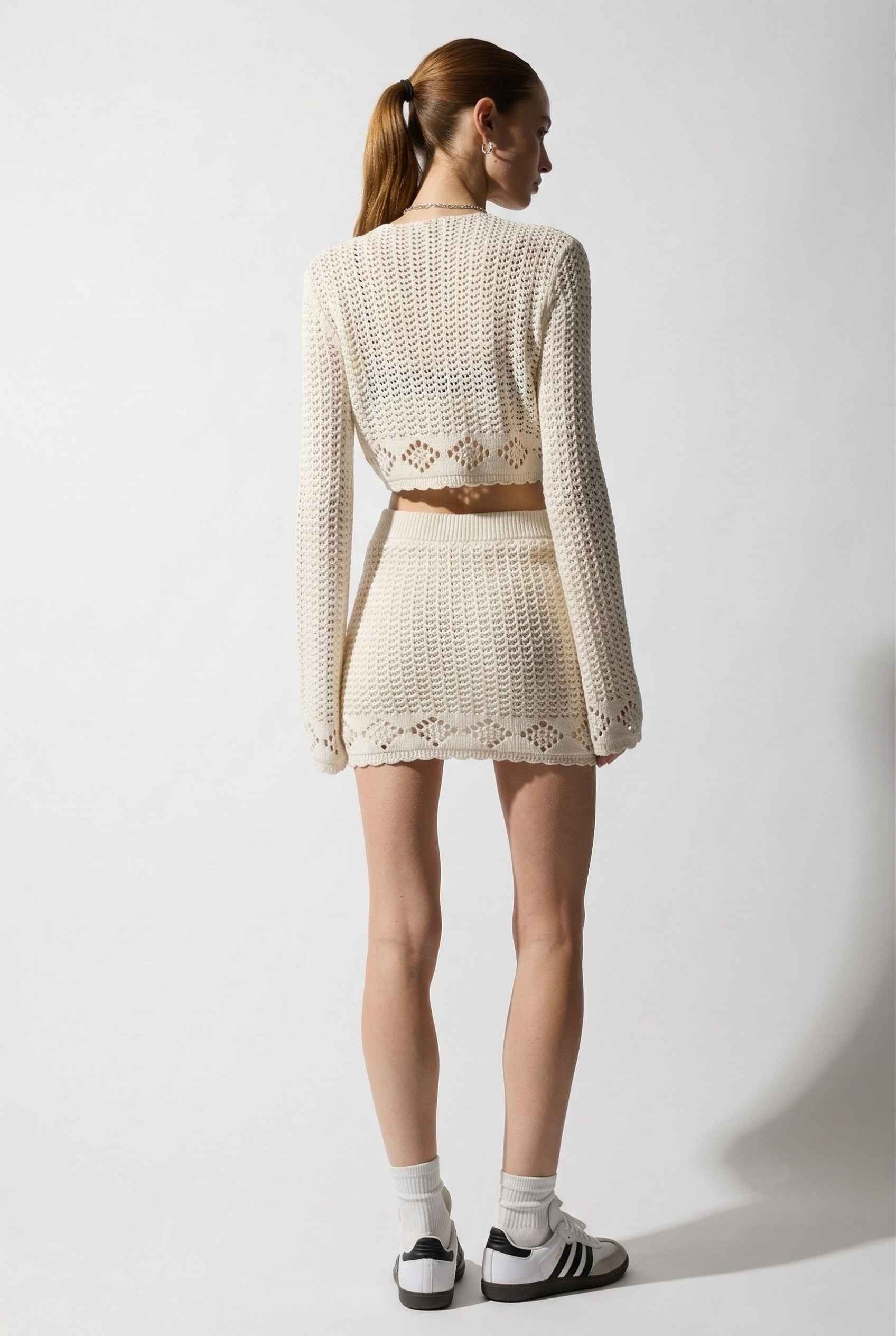 Model wearing Calla Skirt in Ivory Cotton Crochet Mini Skirt with Lettuce Hem paired with matching bolero top and sneakers on a plain background