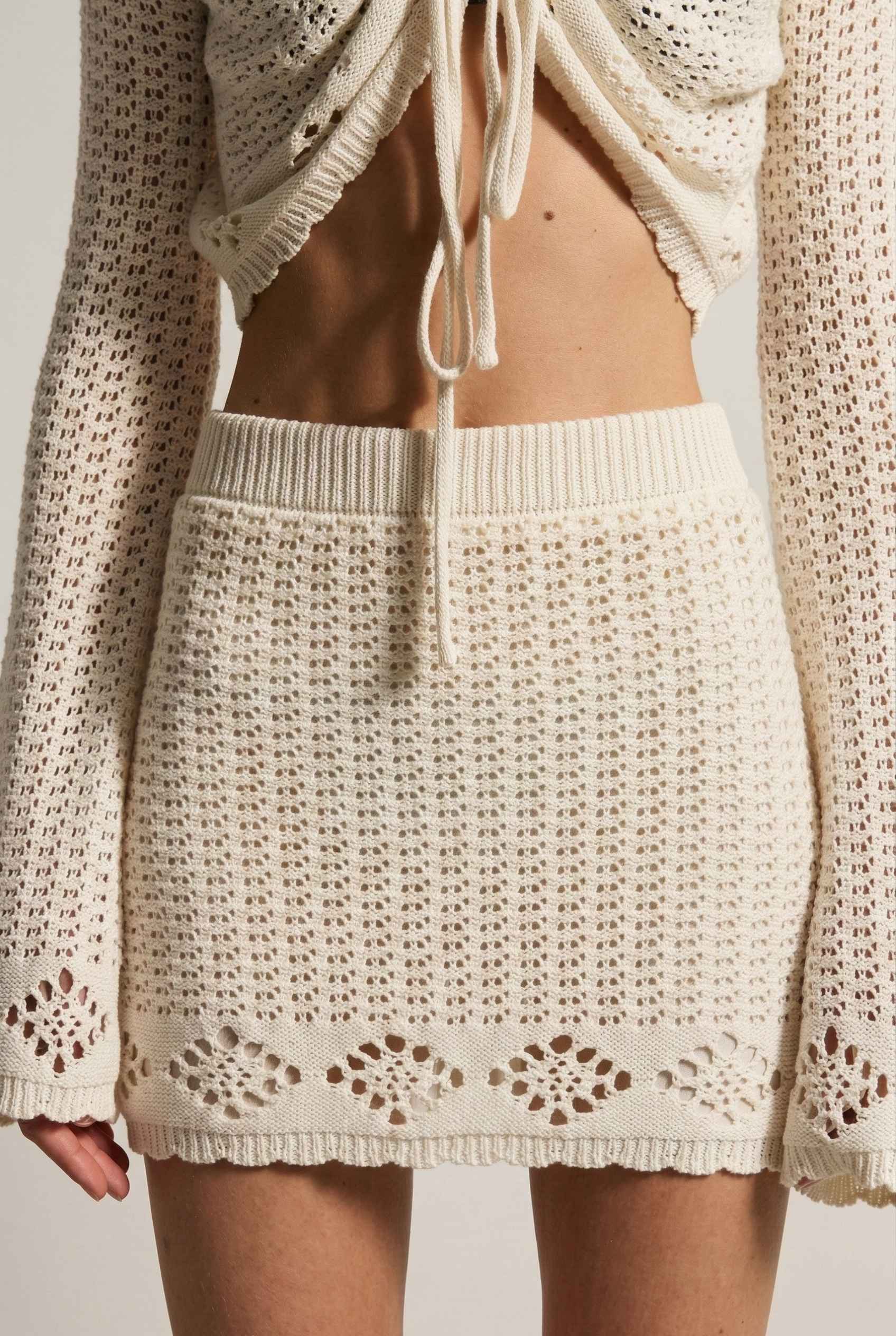 Model wearing Calla Skirt in Ivory Cotton Crochet Mini Skirt with Lettuce Hem paired with matching bolero top on a plain background