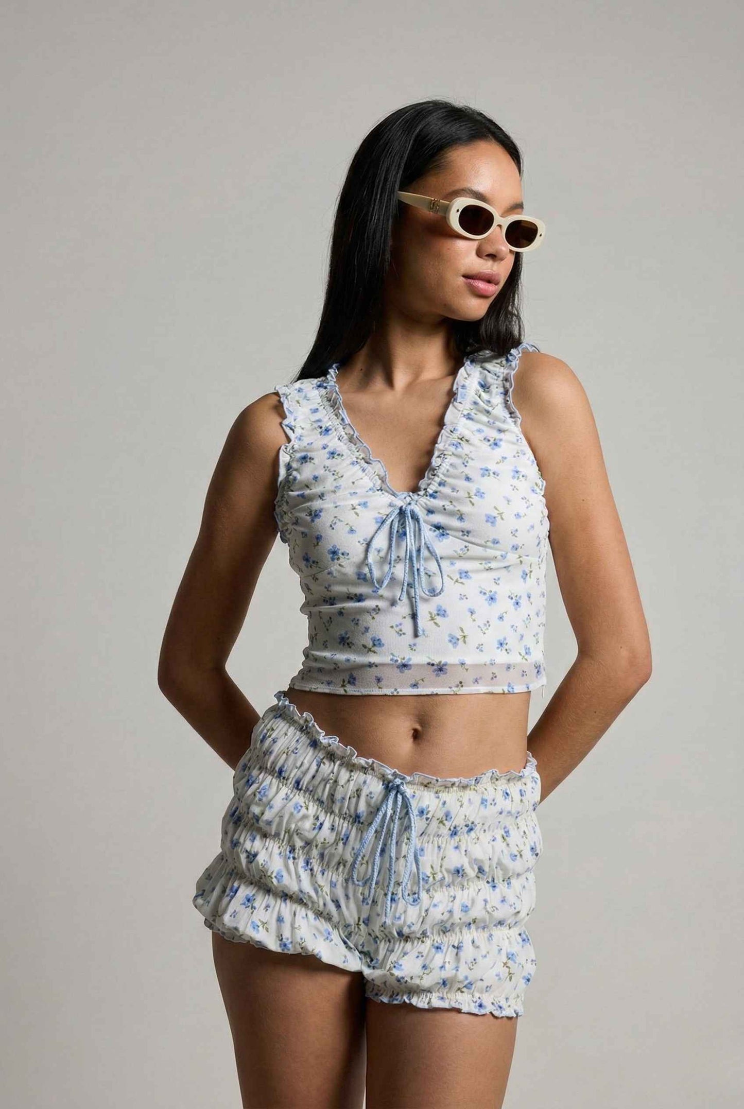 Woman wearing a Callie Top White & Light Blue Floral Ruffle Tie-Front Cami with sunglasses on a plain background