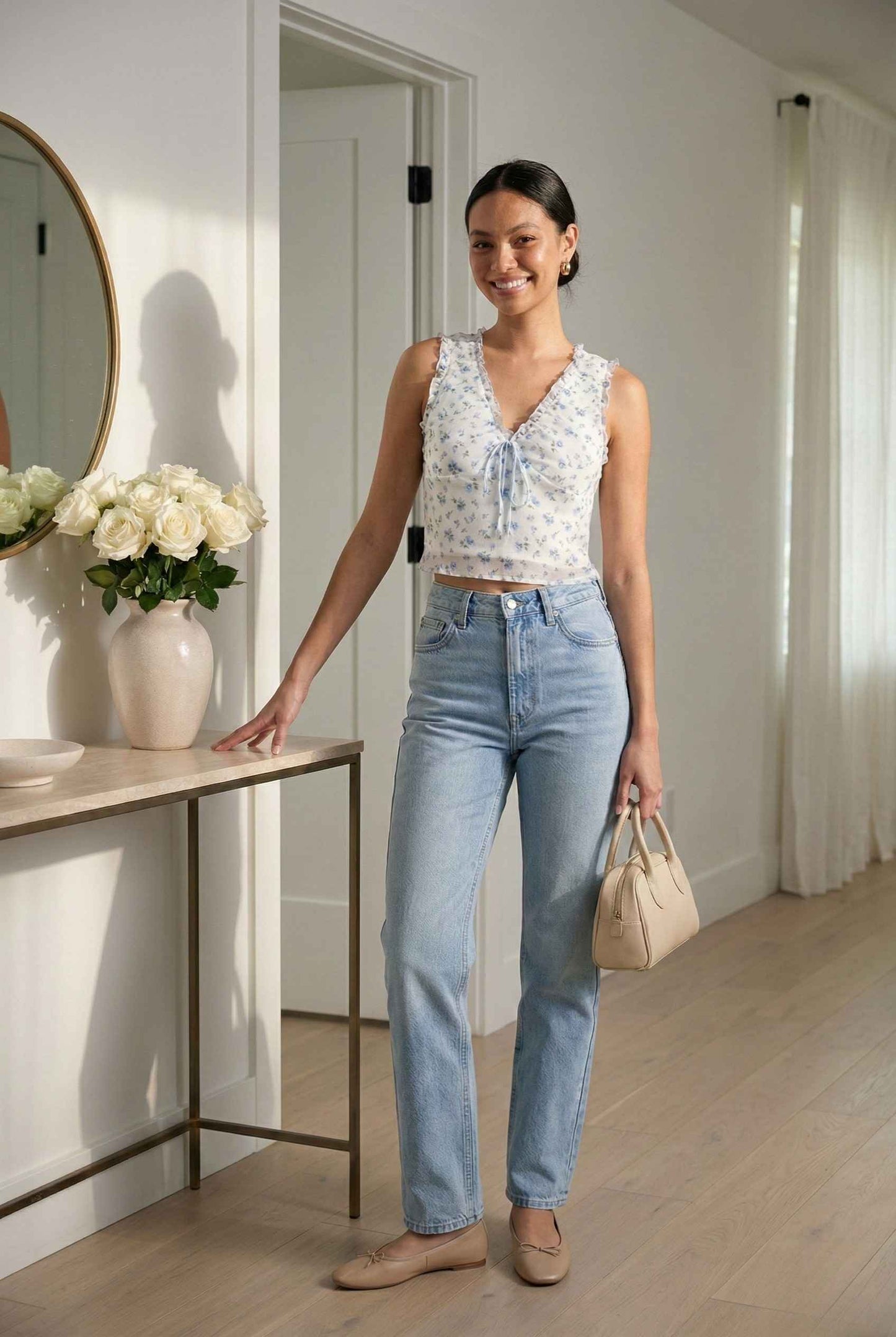 Woman in a Callie Top White & Light Blue Floral Ruffle Tie-Front Cami and blue jeans standing in a room with a table and flowers.